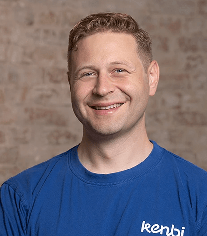 Mann mit kurzem Haar lächelt im blauen T-Shirt vor einer Ziegelwand.