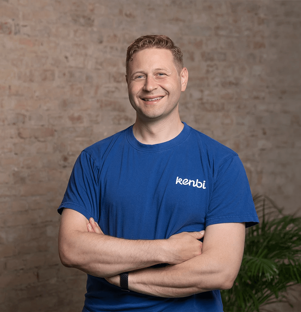 Lächelnder Mann in blauem T-Shirt mit gekreuzten Armen vor Ziegelwand.