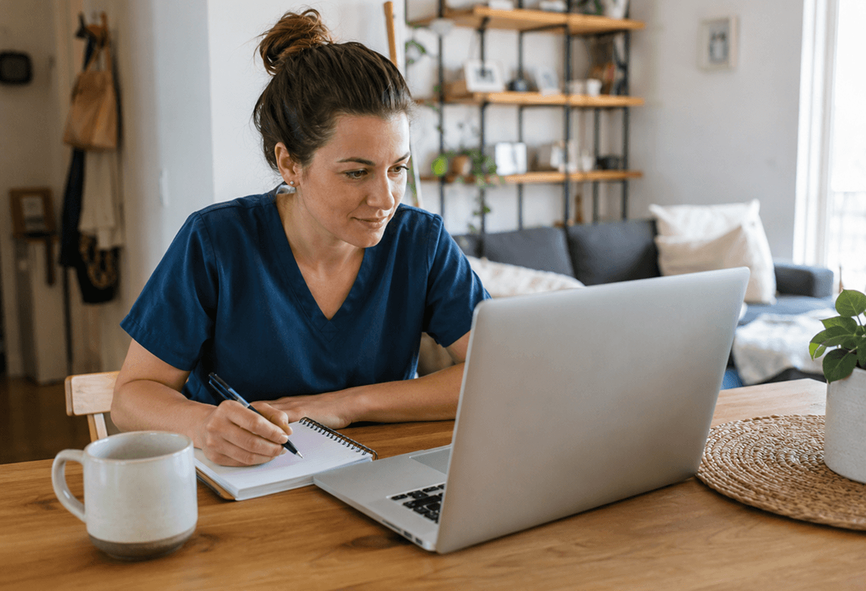 Frau in blauer Kleidung arbeitet an Laptop am Holztisch, schreibt in Notizbuch, gemütliches Wohnzimmer im Hintergrund.