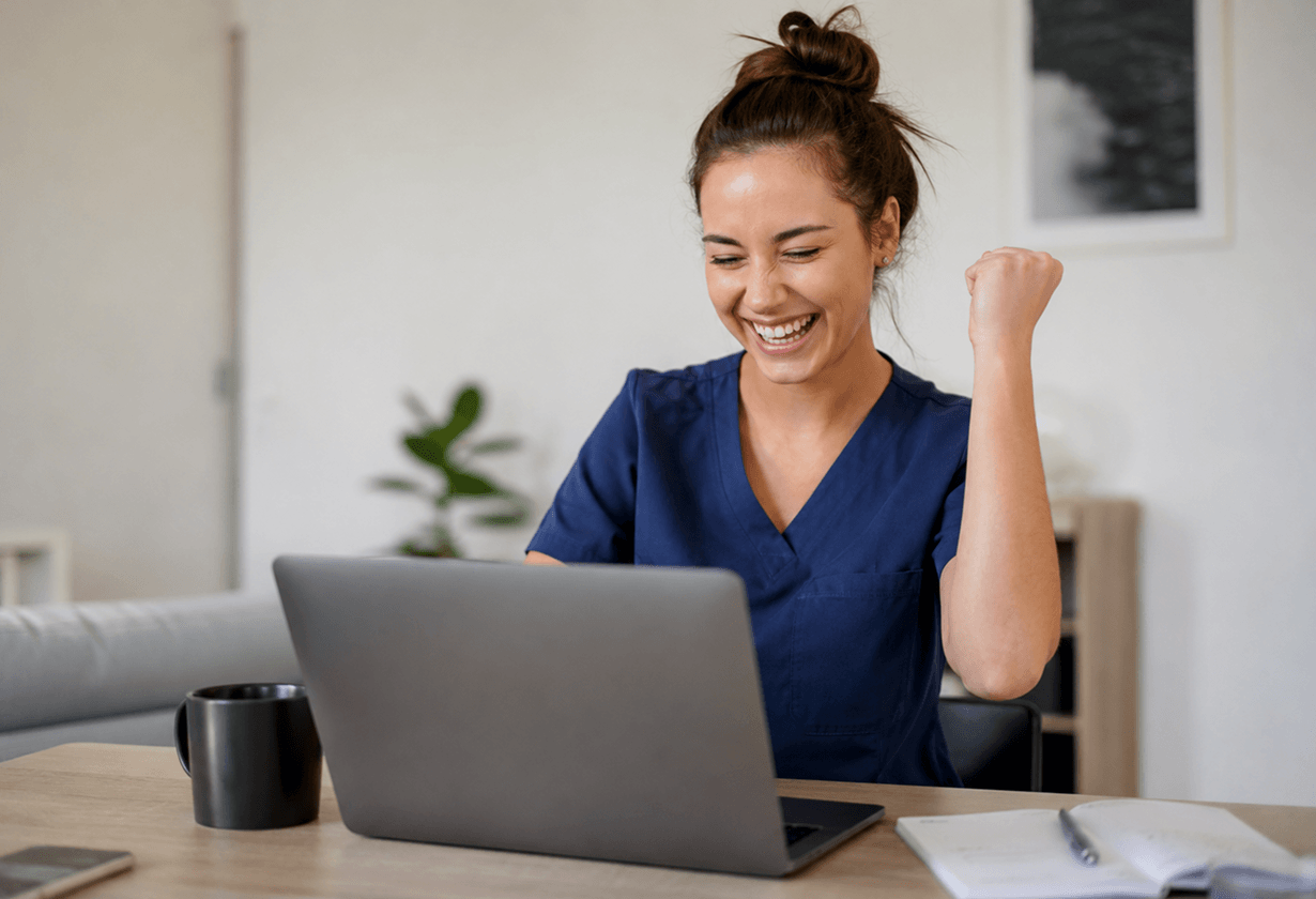 Frau in blauer Kleidung lächelt und jubelt am Laptop, sitzt an Holztisch mit Notizbuch und Kaffeetasse.