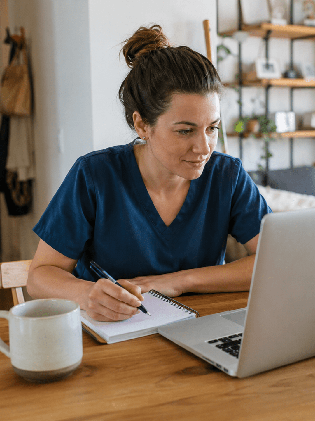 Frau in blauer Arbeitskleidung arbeitet konzentriert am Laptop, macht Notizen, große Tasse im Vordergrund.