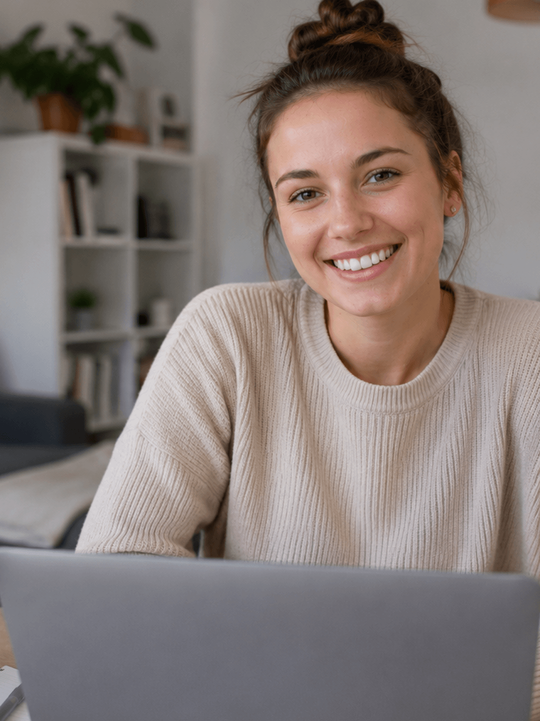 Frau lächelt beim Arbeiten am Laptop, sitzt vor Bücherregal und Pflanze im Hintergrund.