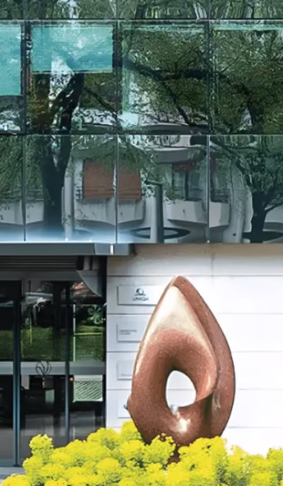 Entrée d'un bâtiment moderne en verre présentant une grande sculpture abstraite brune nichée dans des arbustes à fleurs jaunes.