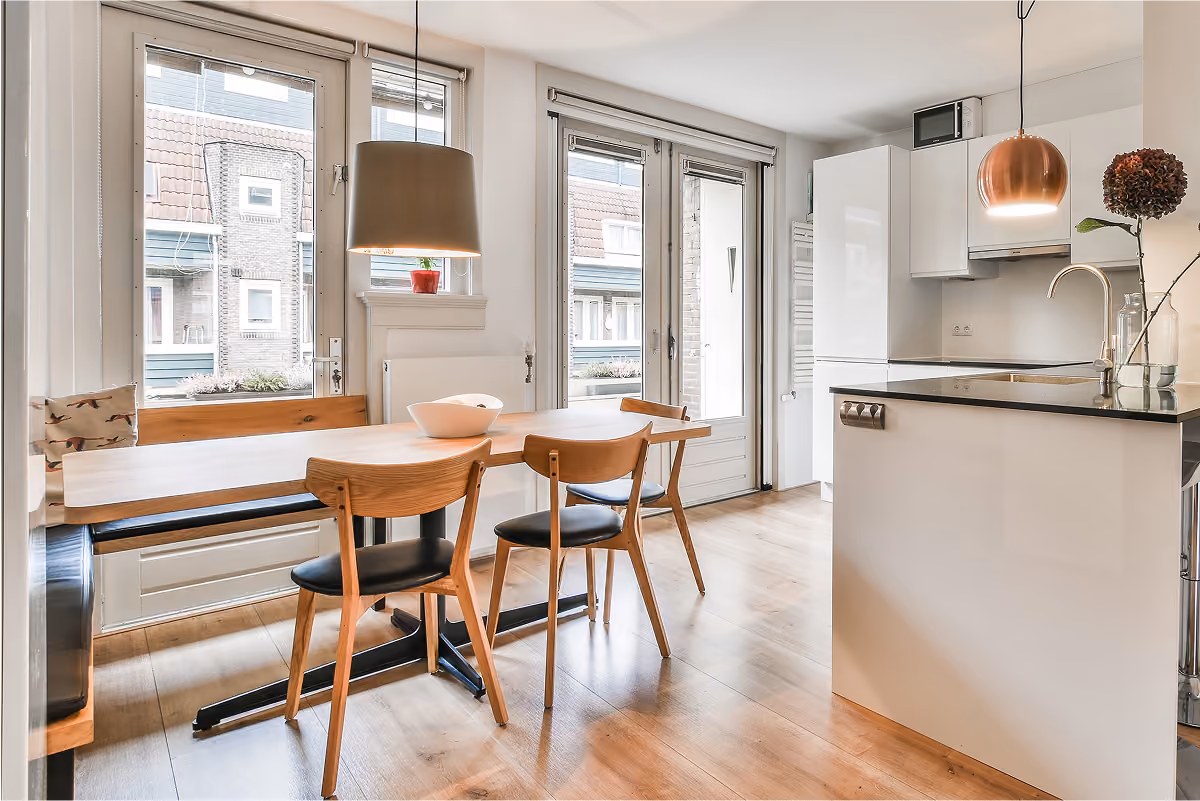 Une salle à manger et cuisine ouverte moderne et lumineuse, dotée d'une table à manger en bois avec des chaises, d'un îlot de cuisine blanc et d'une suspension en cuivre.