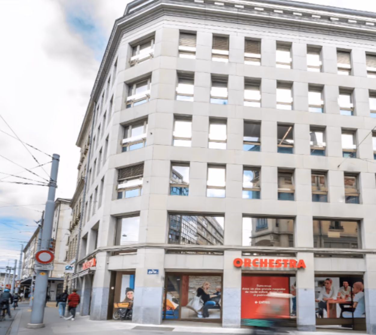 Vue d'angle d'un bâtiment urbain en pierre à plusieurs étages avec une devanture de magasin Orchestra au rez-de-chaussée et des lignes aériennes de tramway.