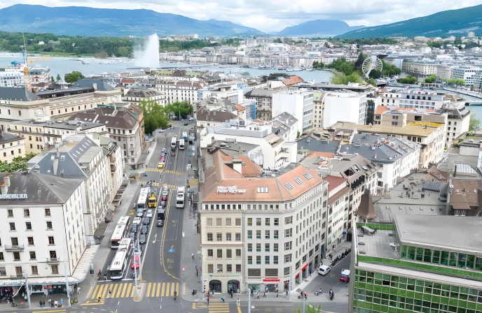 Large vue aérienne du paysage urbain de Genève, s'étendant jusqu'au lac Léman avec le Jet d'Eau et les montagnes environnantes en arrière-plan.