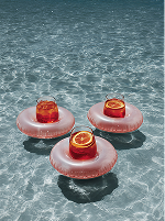 Three orange drinks in round pink floaters floating on clear swimming pool water.