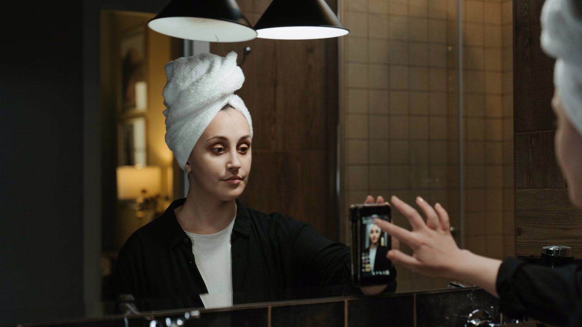 Woman in a hair towel filming herself on a smartphone in a bathroom mirror