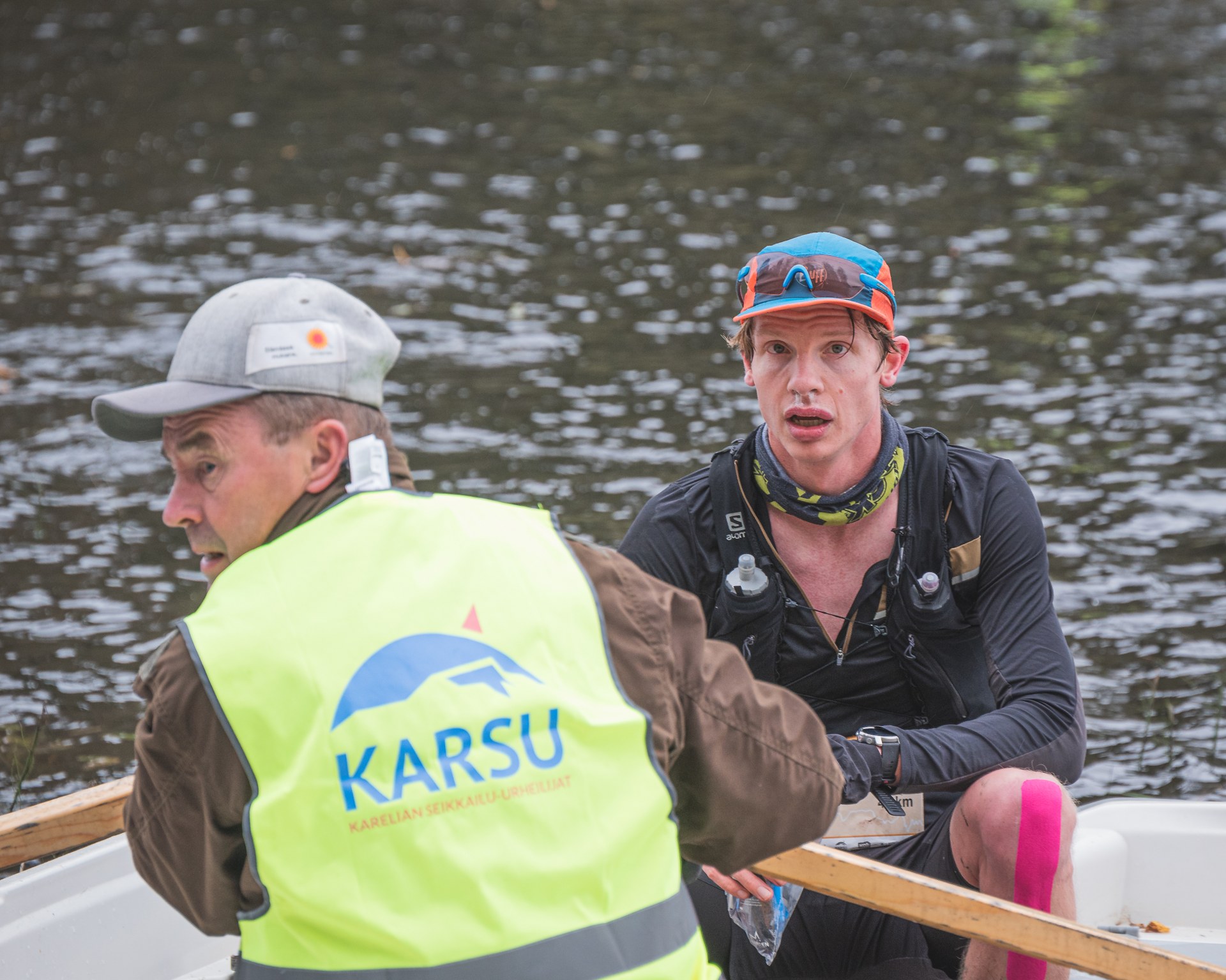 Two men in a rowboat on water; the man facing forward wears a black outfit, orange and blue cap, and pink kinesiology tape on his knee, while the man facing backward wears a gray cap and a yellow safety vest with 'KARSU' written on the back.