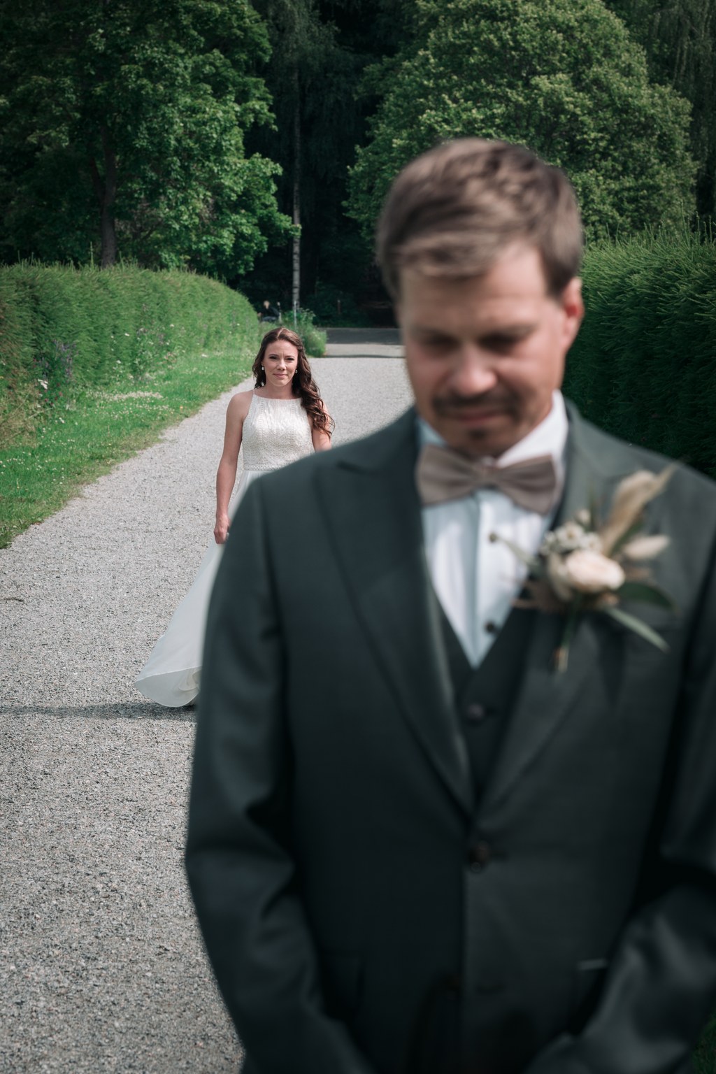 Morsian valkoisessa mekossa kävelee sorapolkua kohti sulhasta tummassa puvussa rusetti ja boutonniere-puvussa vihreiden puiden ja pensaiden ympäröimänä.