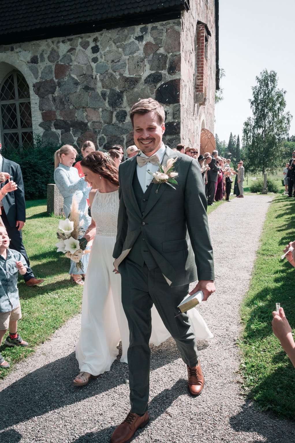 Hymyilevä sulhanen vihreässä puvussa ja ruskeissa kengissä kävelee sorapolulla valkoiseen mekkoon pukeutuneen morsiamen vieressä, joka pitelee kimppua. Häävieraat seisovat polun varrella kivirakennuksen ja vihreiden puiden lähellä kirkkaan taivaan alla.