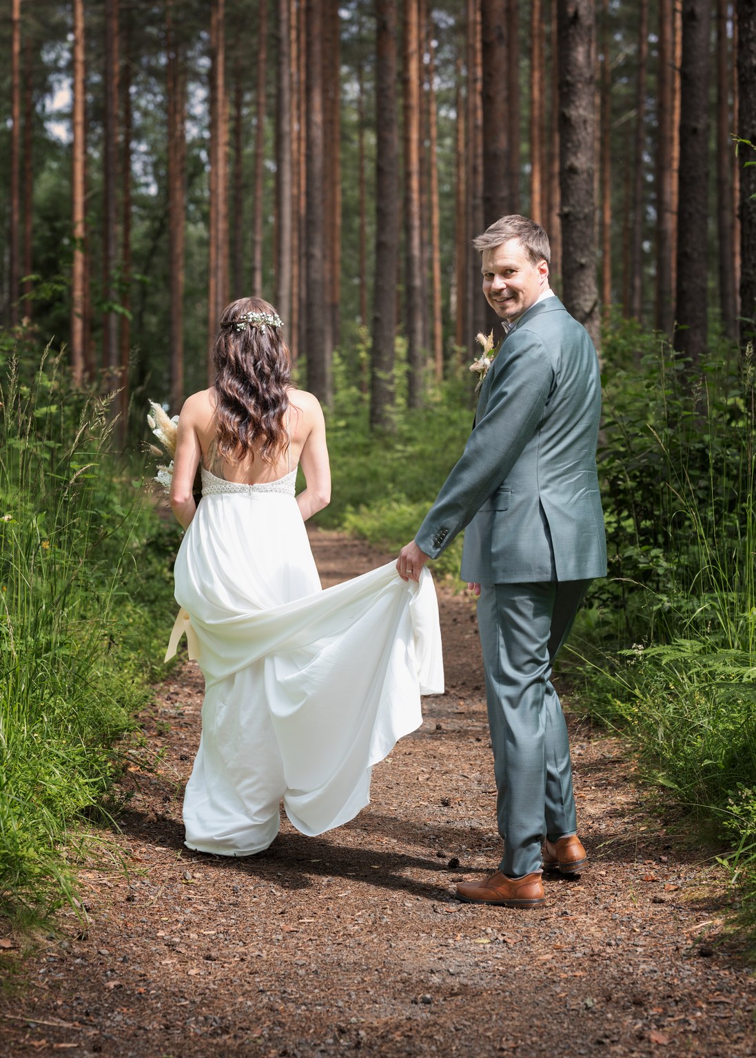 Morsian ja sulhanen kävelevät metsäpolulla korkeiden puiden kanssa; vaaleansinisessä puvussa oleva sulhanen pitää morsiamen valkoista pukujunaa kävellessä eteenpäin kimpussa.