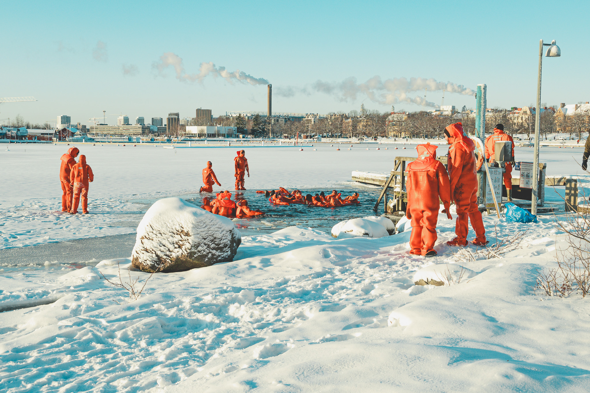 Group of people in orange survival suits practicing cold water rescue or winter swimming in a snowy, icy outdoor setting near a dock with cityscape and smoke stacks in the background.
