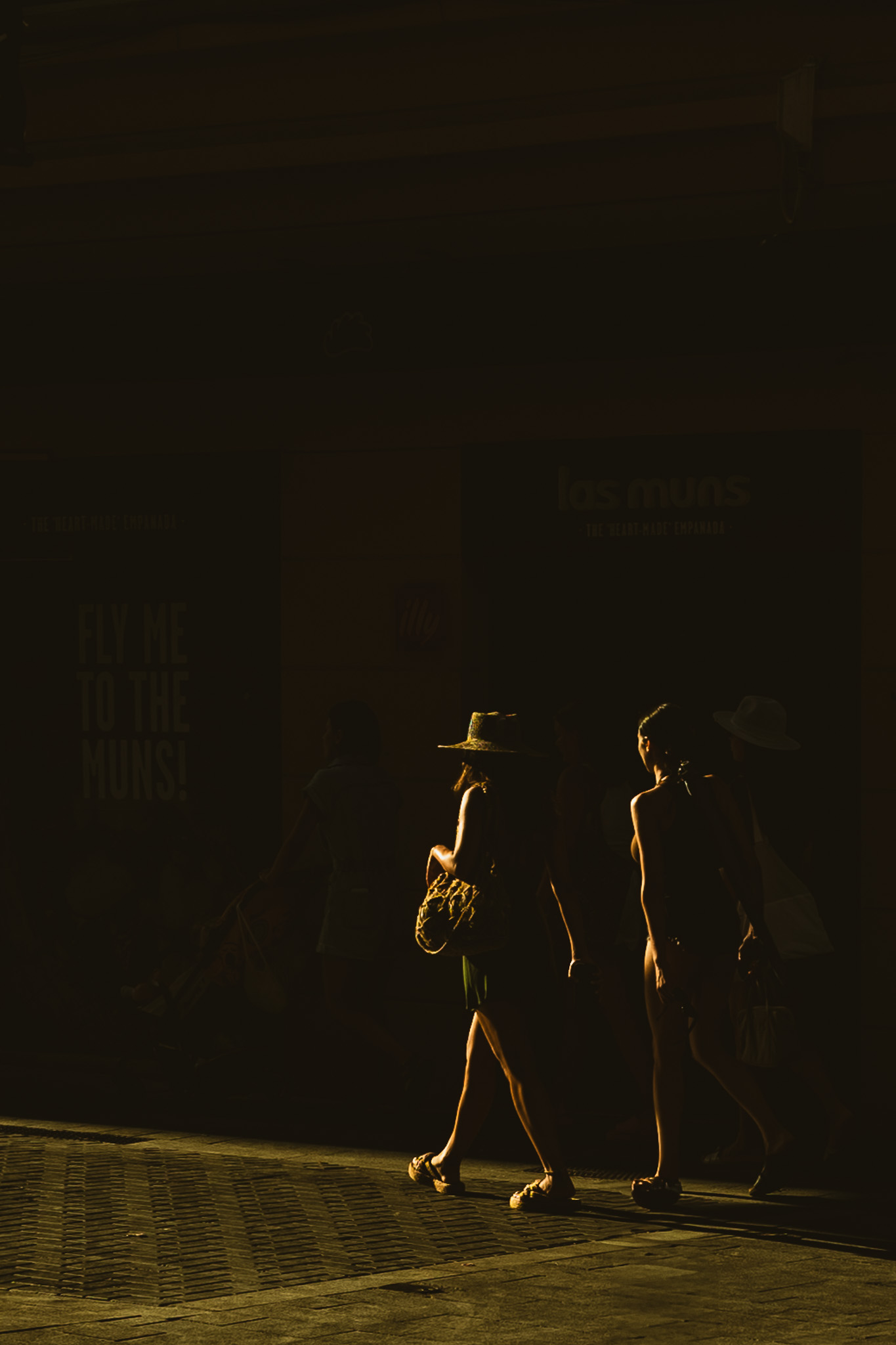 Silhouetted group of four people walking on a sunlit street, two women wearing hats and carrying bags, with strong contrast of light and shadow.