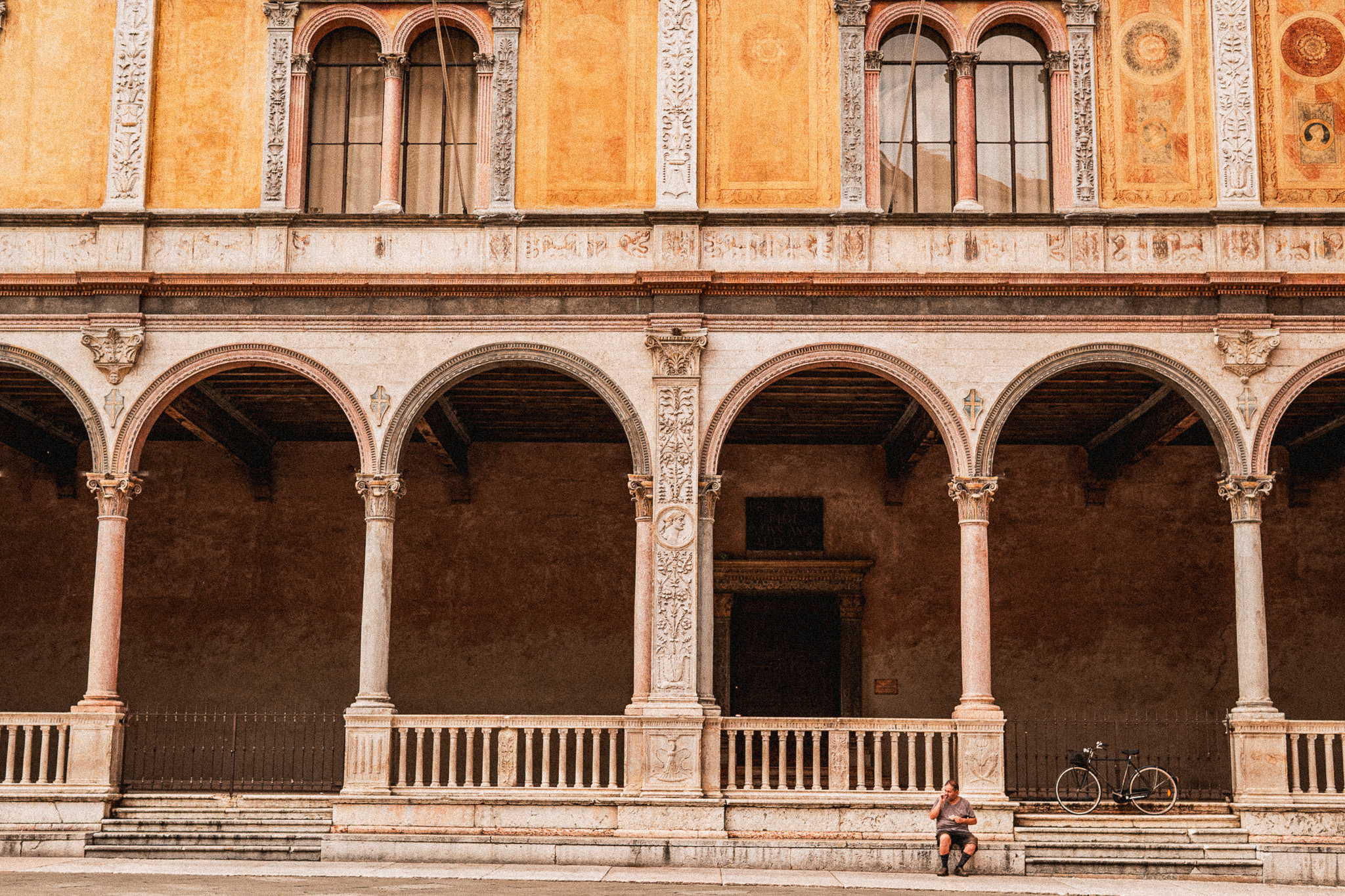 Historic building facade with tall, ornate arches and columns in warm yellow and beige tones, featuring intricate carvings and decorative windows above; a man sits on the stone steps near a black bicycle leaning against the railing.