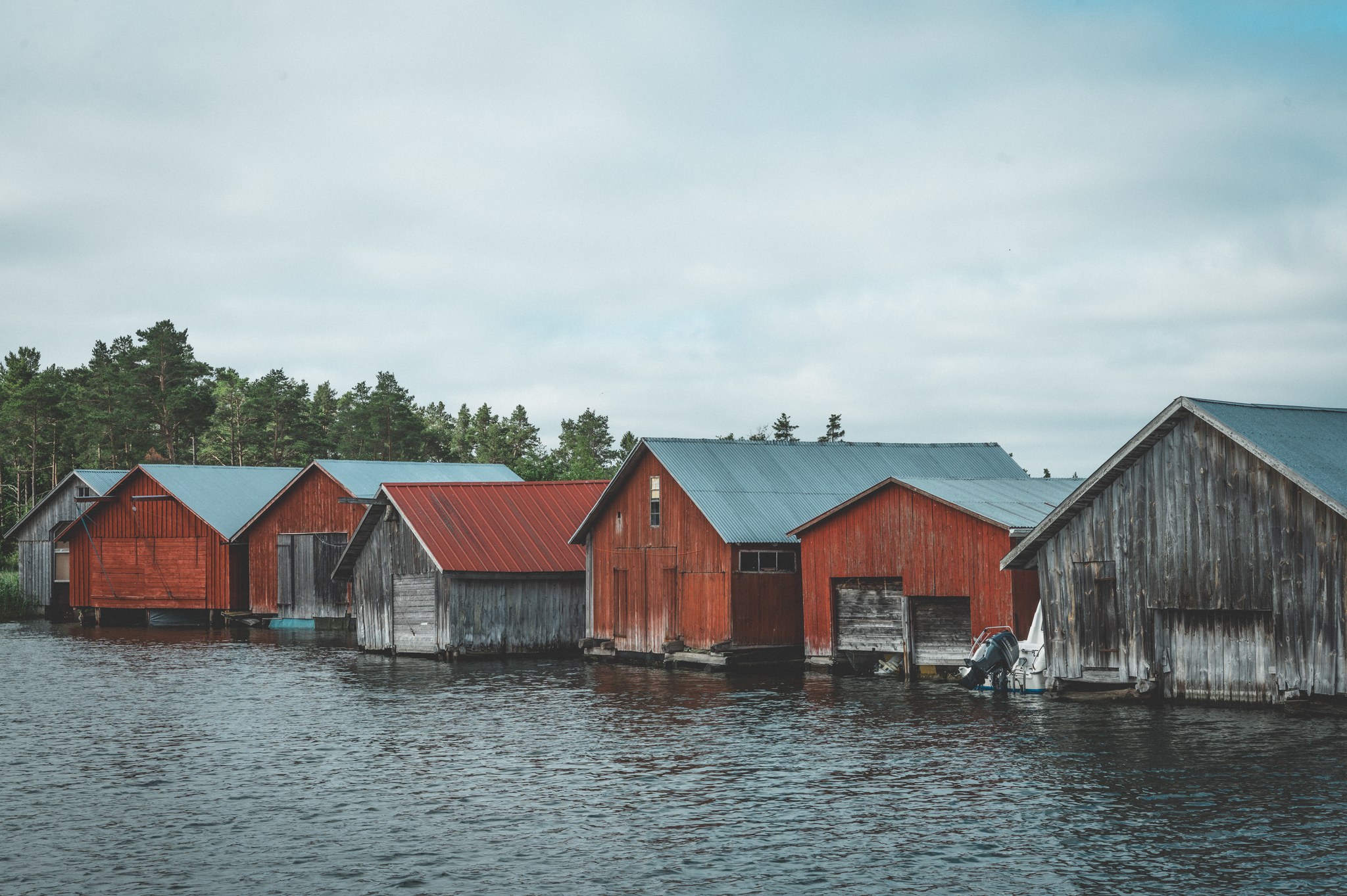 Row of rustic boathouses with red and gray wooden siding and metal roofs lined up over calm water with a forest in the background under a cloudy sky.