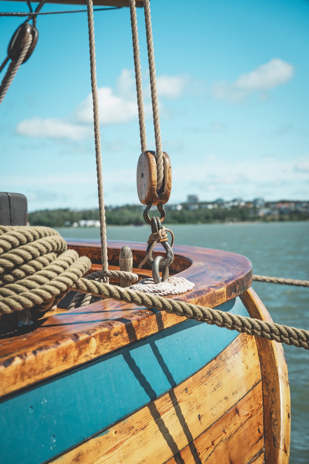 Close-up of a wooden sailboat's bow with coiled ropes, a wooden pulley block hanging from rigging, and a calm water body with a distant shoreline under a blue sky with white clouds.