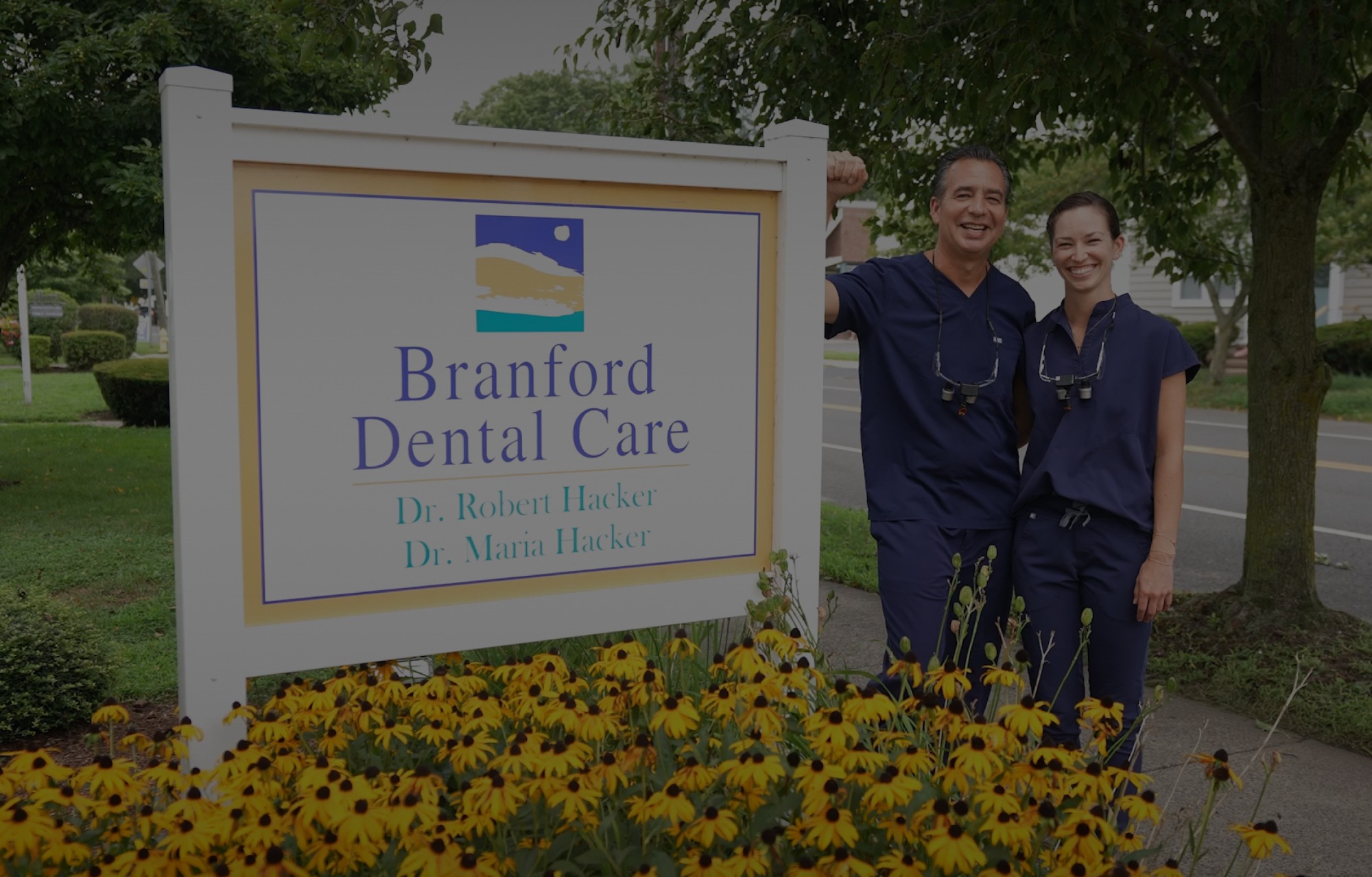 Two dentists in blue scrubs standing by a sign for Branford Dental Care with yellow flowers in front.