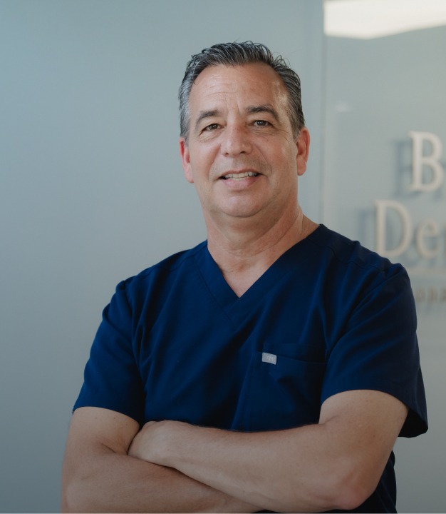 Smiling male healthcare professional in navy scrubs with arms crossed standing indoors.