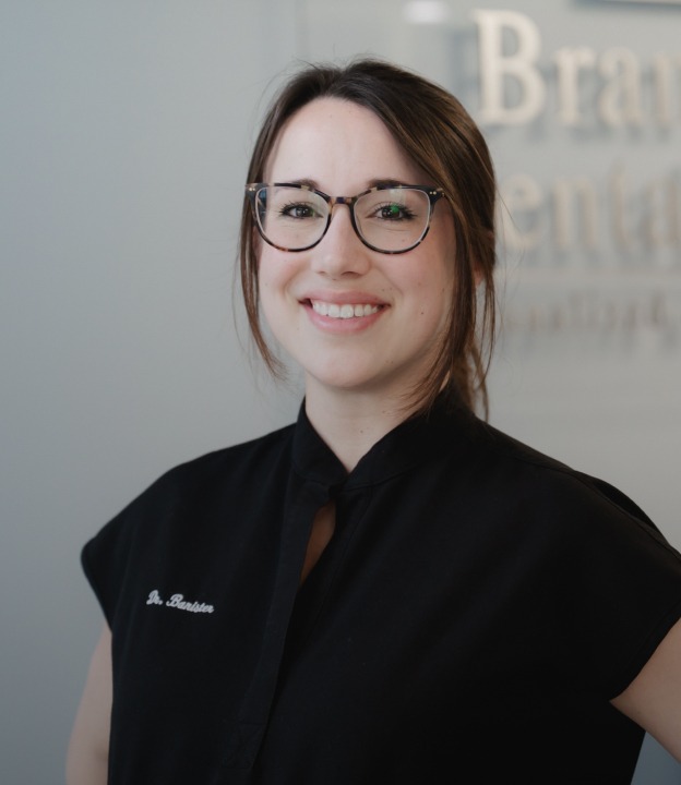 Smiling woman with glasses in black medical attire standing in front of a blurred dental office sign.
