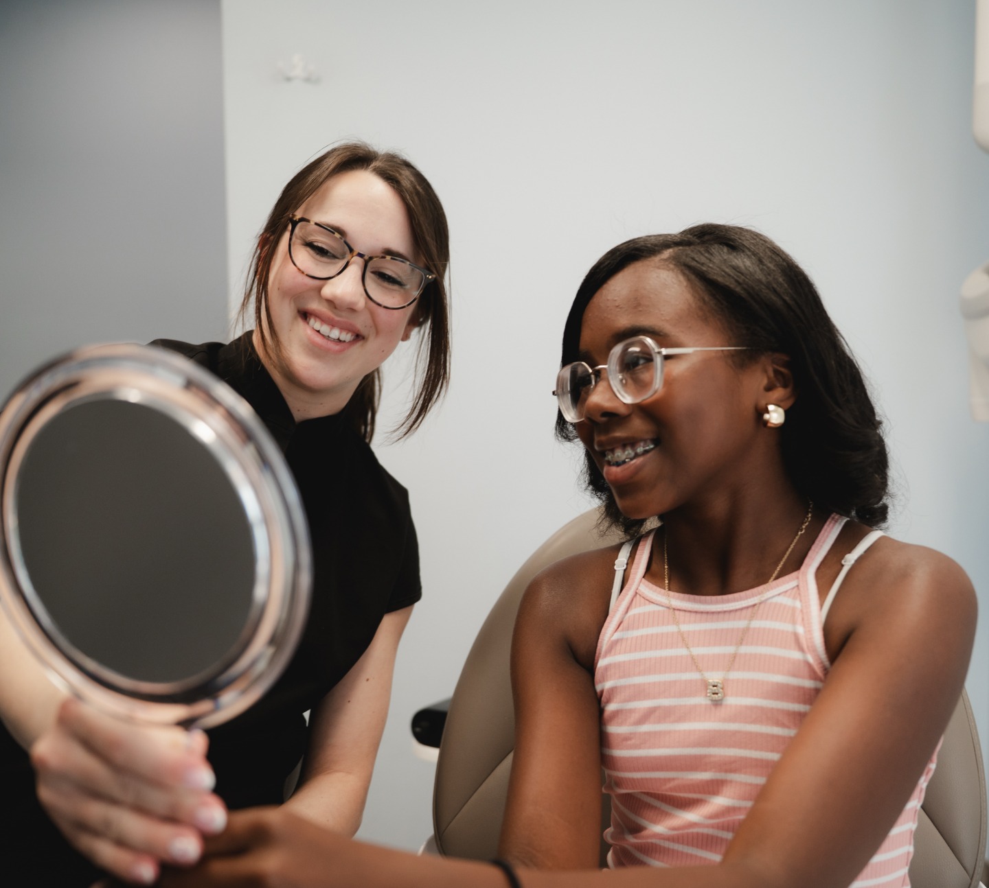 Woman shows a smiling girl with braces a hand mirror while sitting in a chair in a light room.