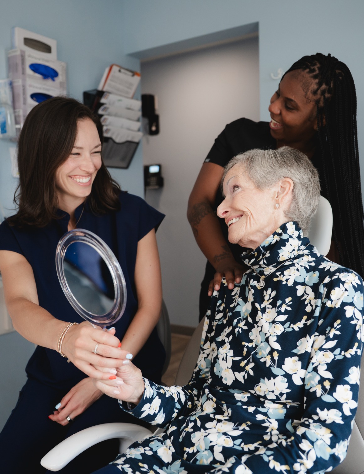 An elderly woman in a floral top holds a mirror, smiling with two smiling women in a medical setting.