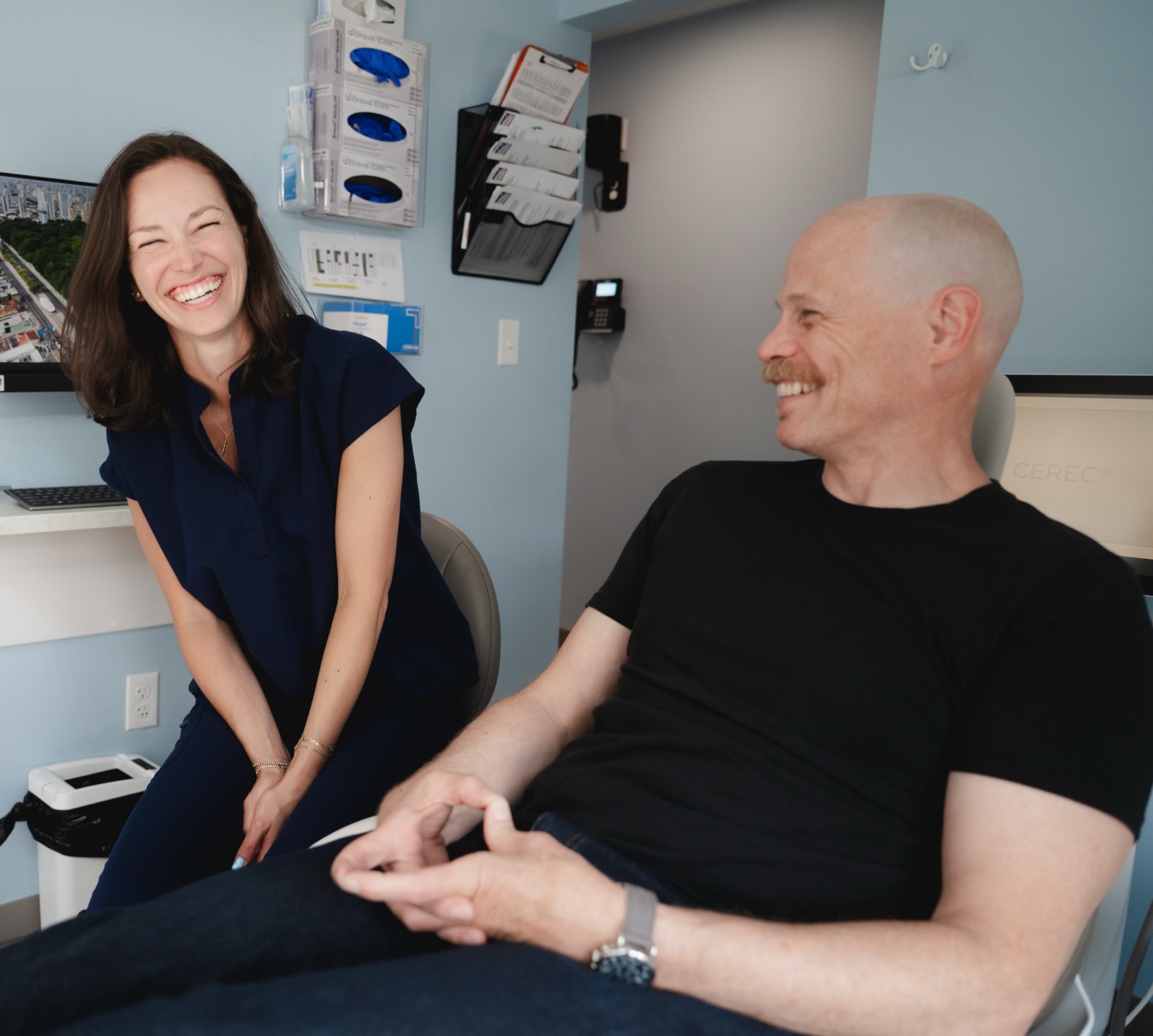 Man with mustache sits smiling at a laughing woman in a navy shirt in a light blue office room.