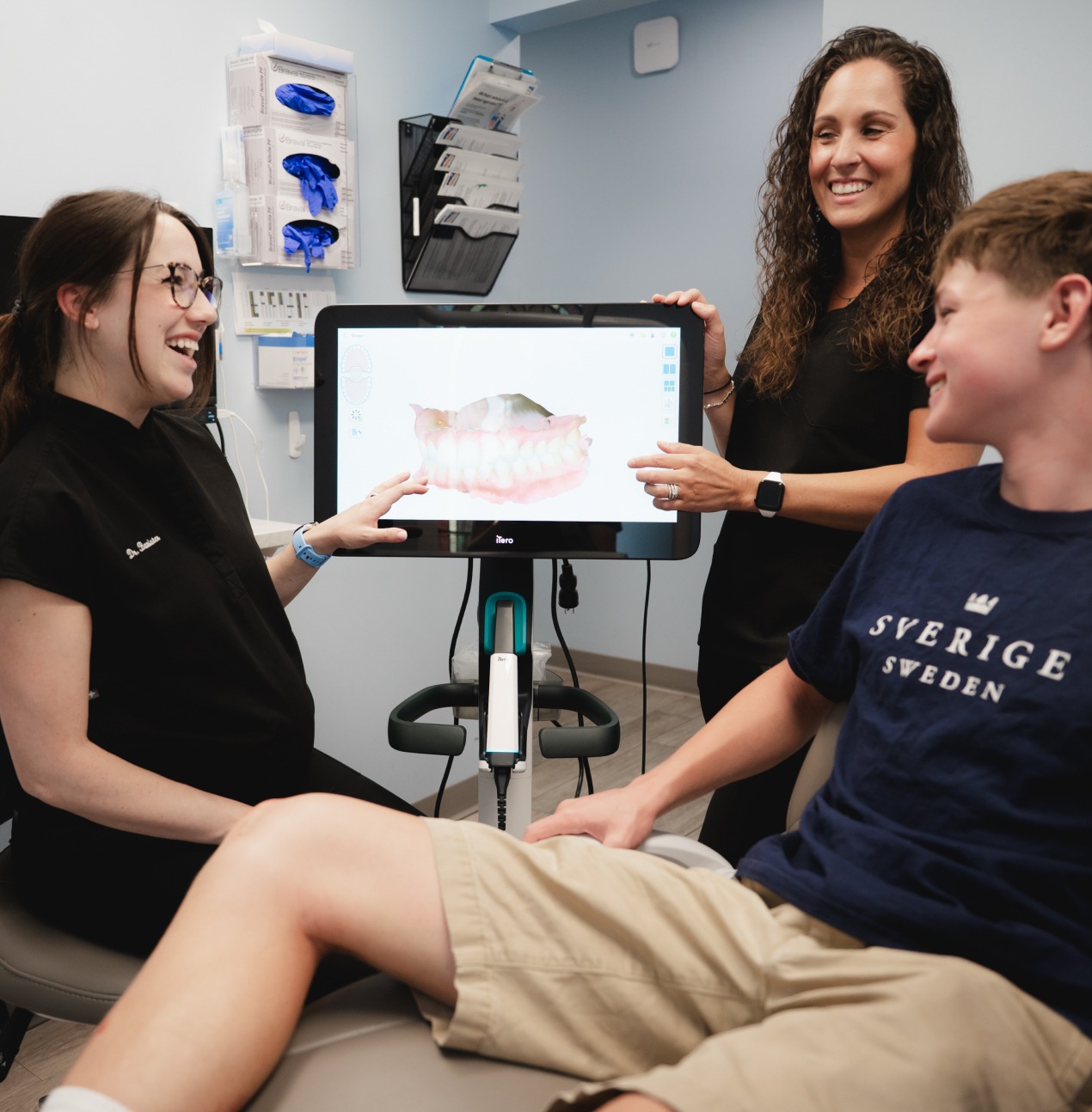 Two dental professionals show a 3D teeth model on a screen to a smiling boy in a dental office.