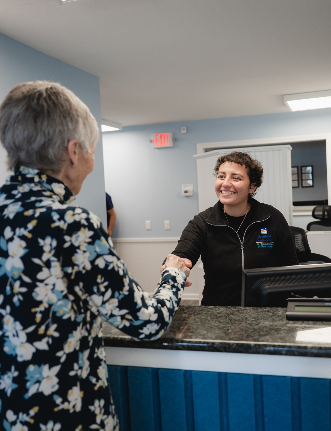 Woman receptionist smiling and shaking hands with an older woman at a dental office counter.
