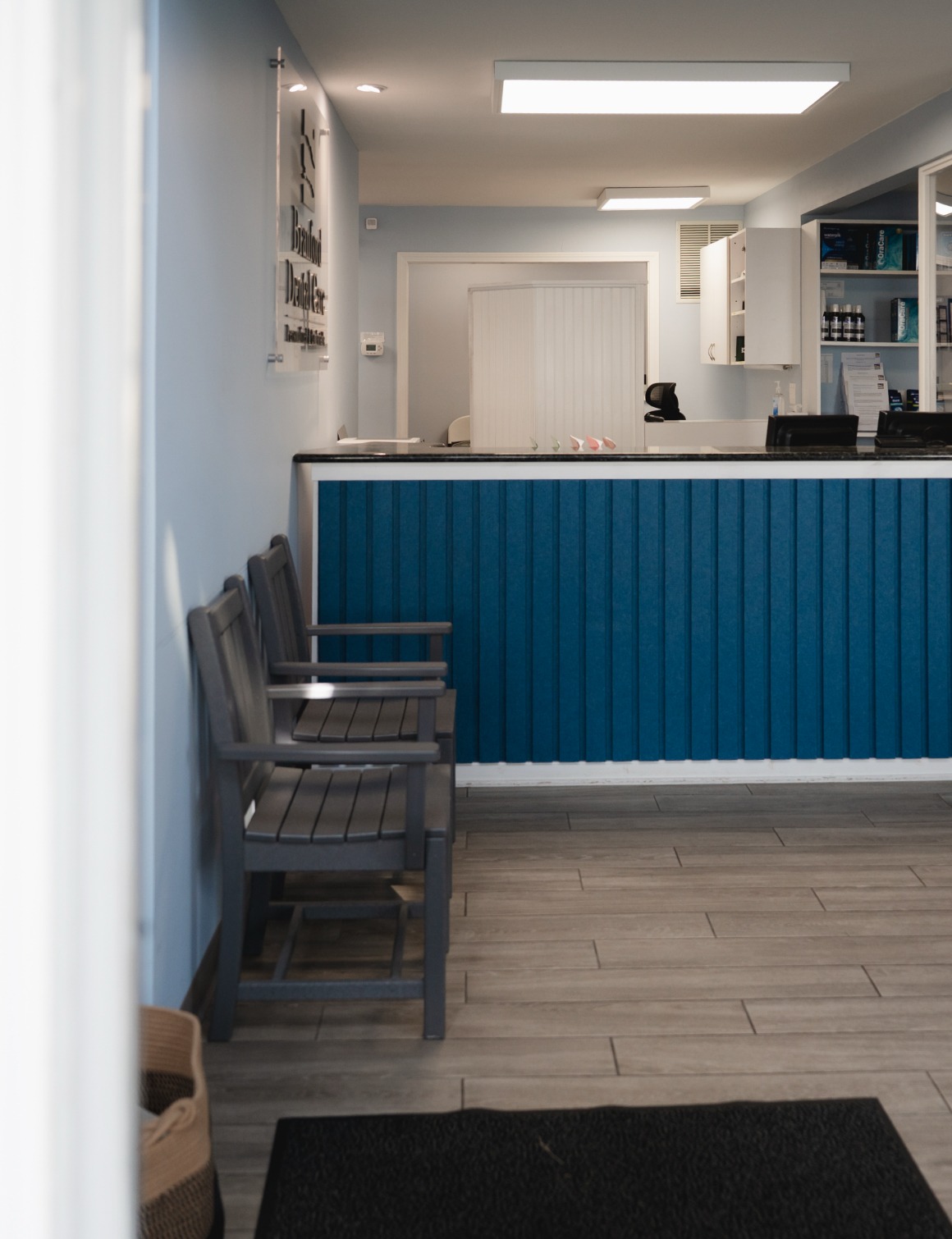 Dental office waiting area with gray chairs and blue front desk under fluorescent lights.