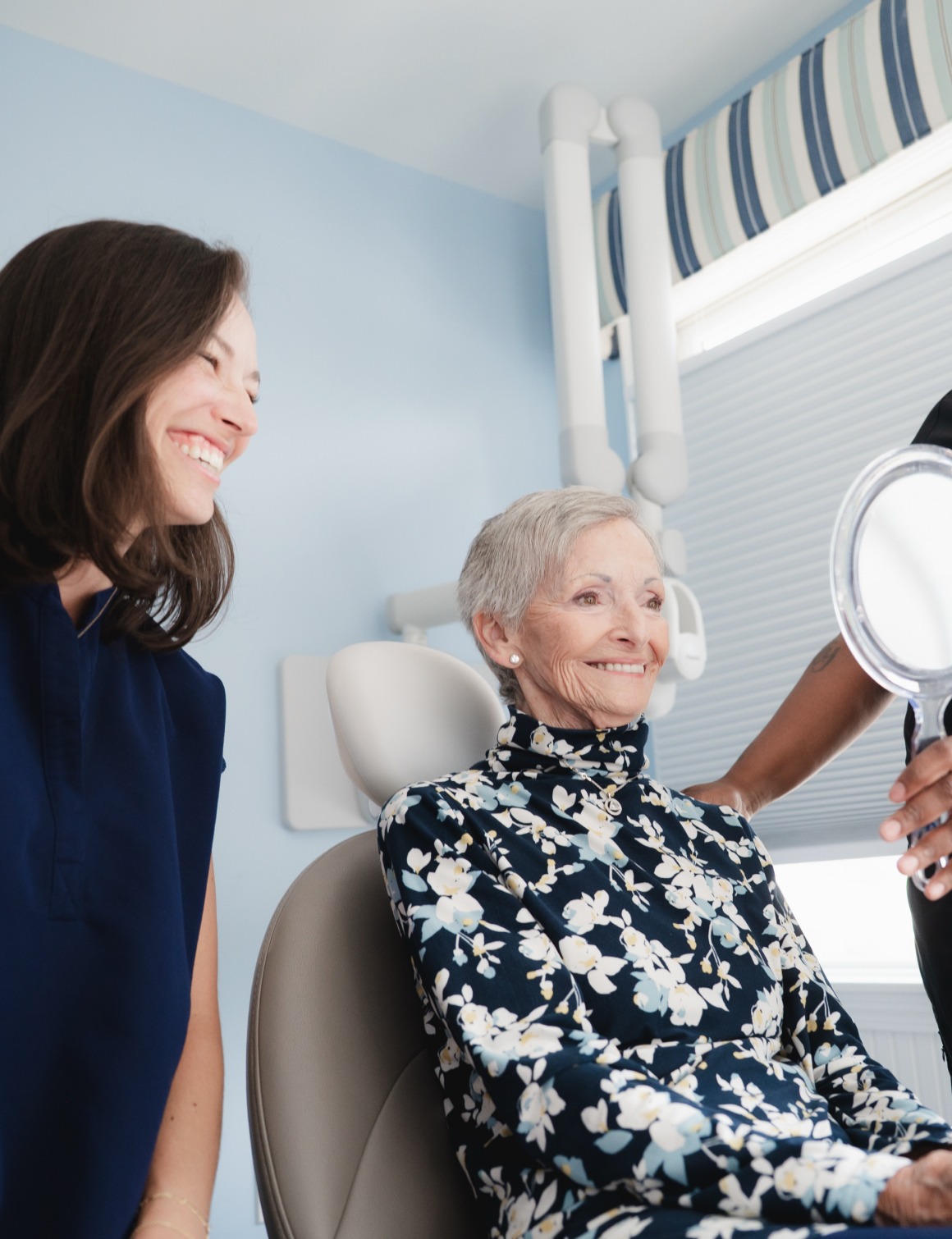 Elderly woman in floral dress sitting in dental chair smiling, dentist holds mirror, assistant nearby.