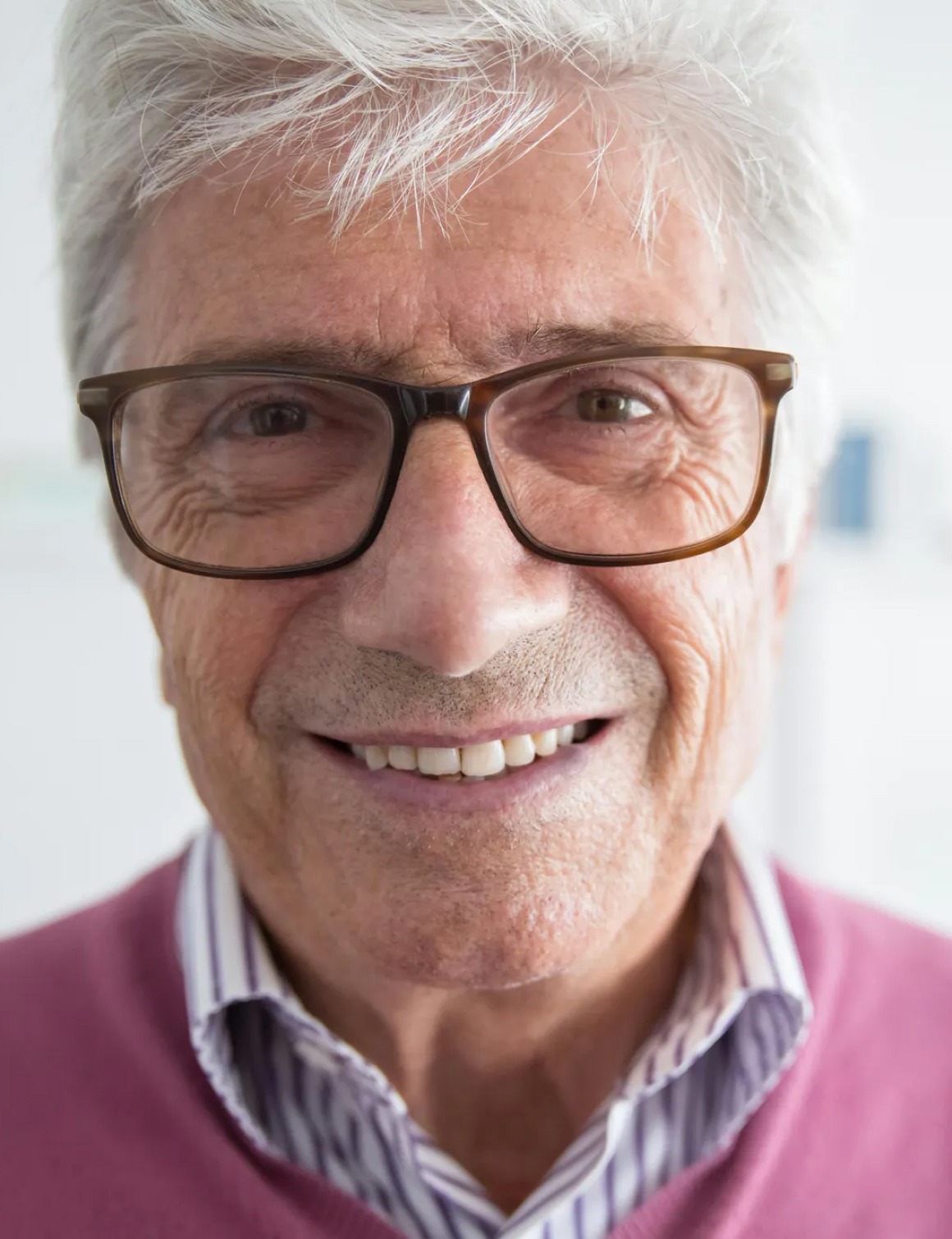 Close-up of an elderly man smiling, wearing glasses and a pink sweater over a striped shirt.