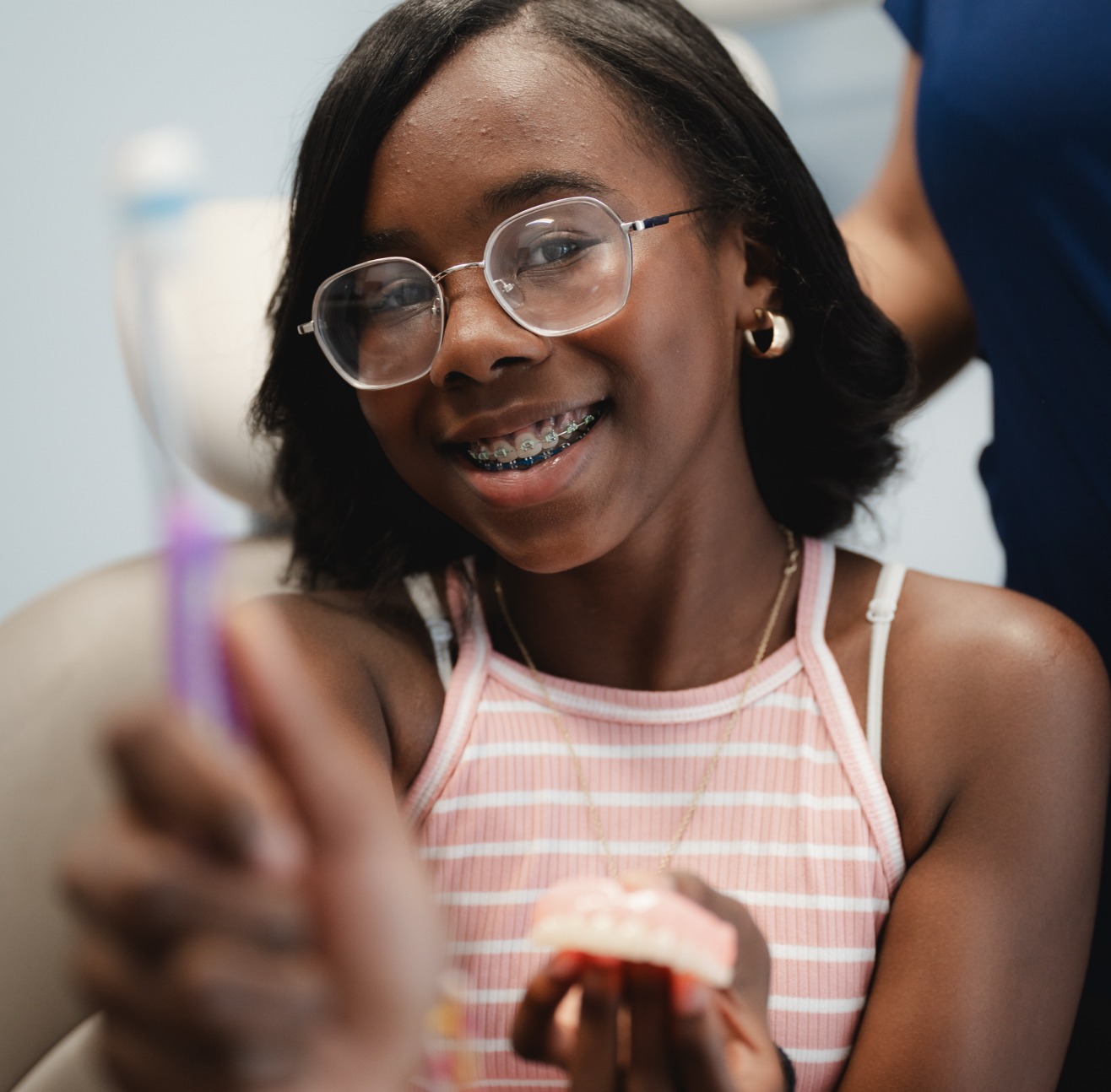 Smiling girl with braces and glasses holding dental model and toothbrush in a dental office.