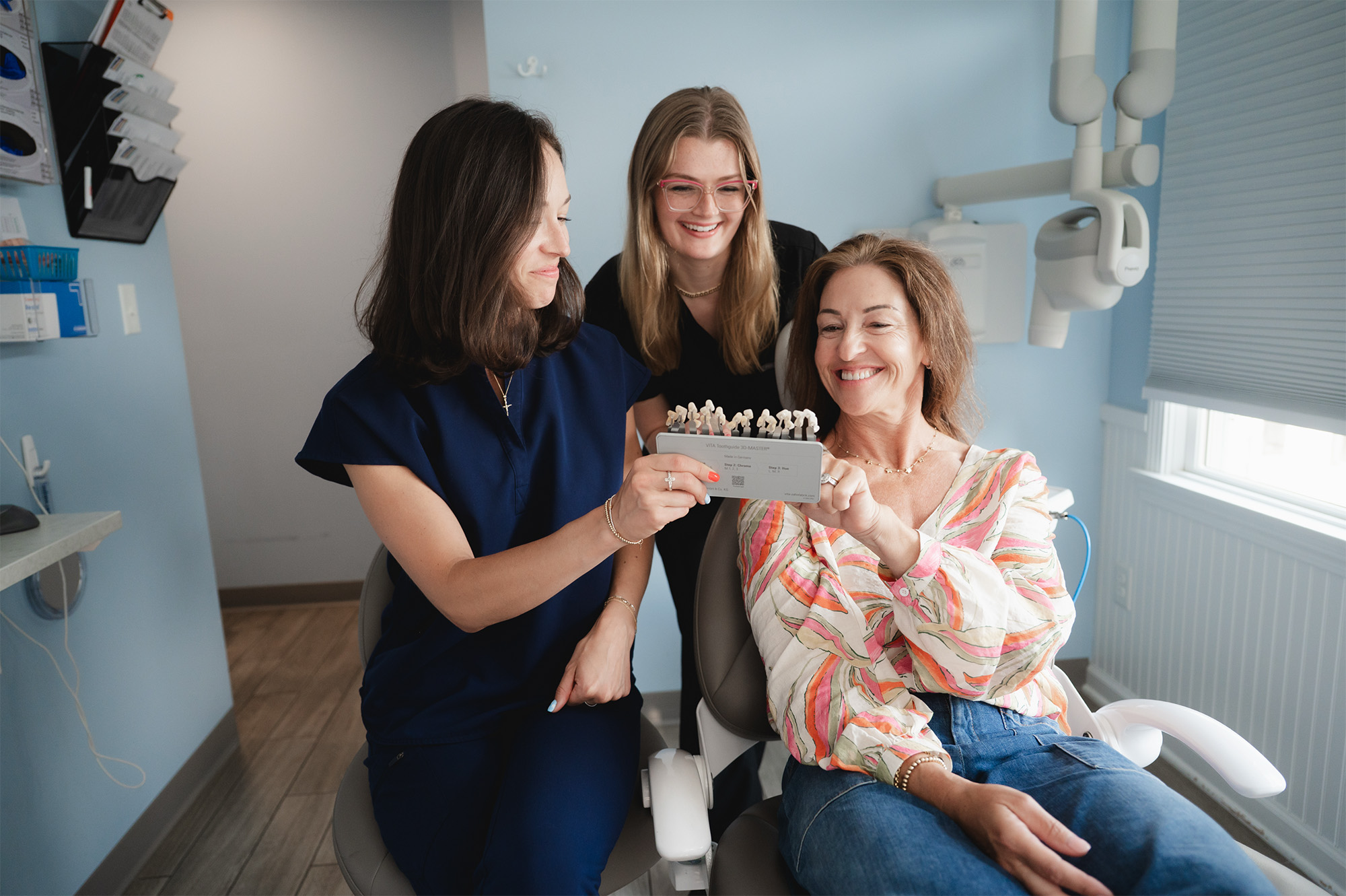 Three women in a dental office examining a dental shade guide together, smiling and seated.