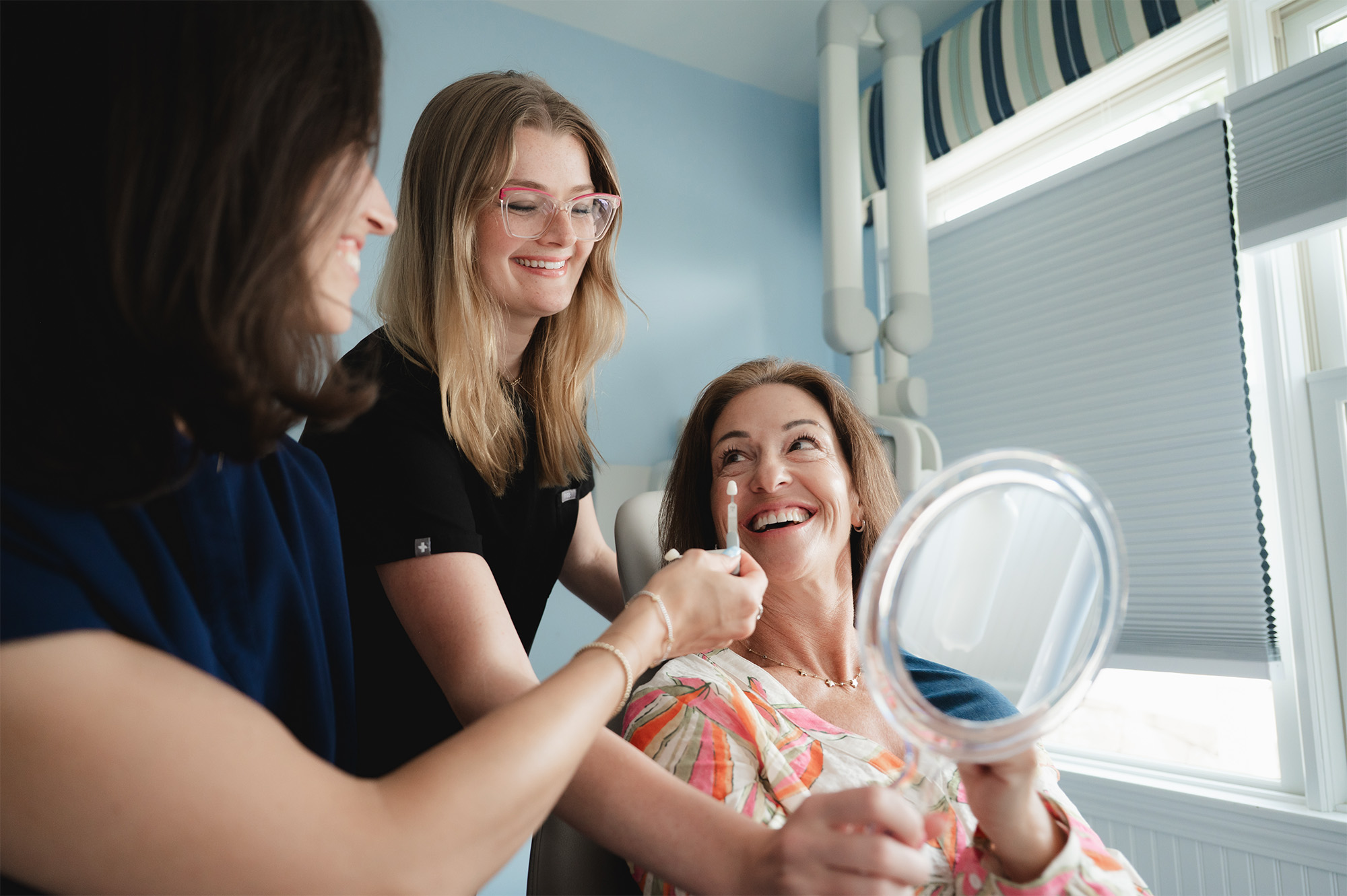 Woman holding mirror smiles as two dental professionals show a shade guide for teeth color matching.