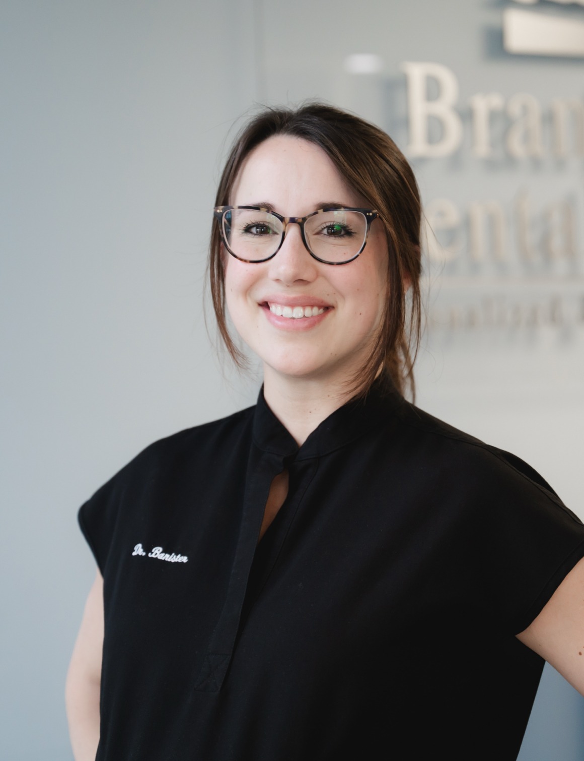 Smiling woman with glasses wearing a black shirt embroidered with 'Dr. Banister' in an office.