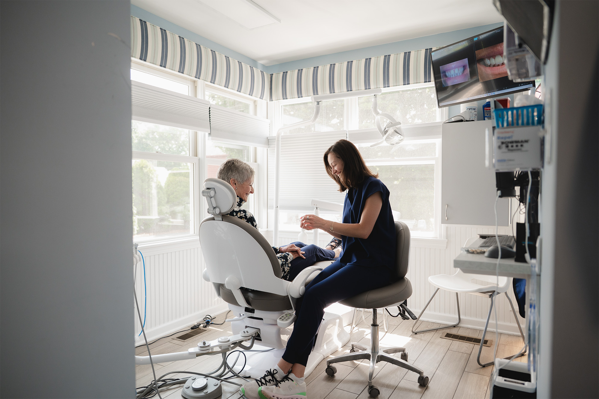 Dentist smiling and talking with an older woman seated in dental chair in a sunny room.