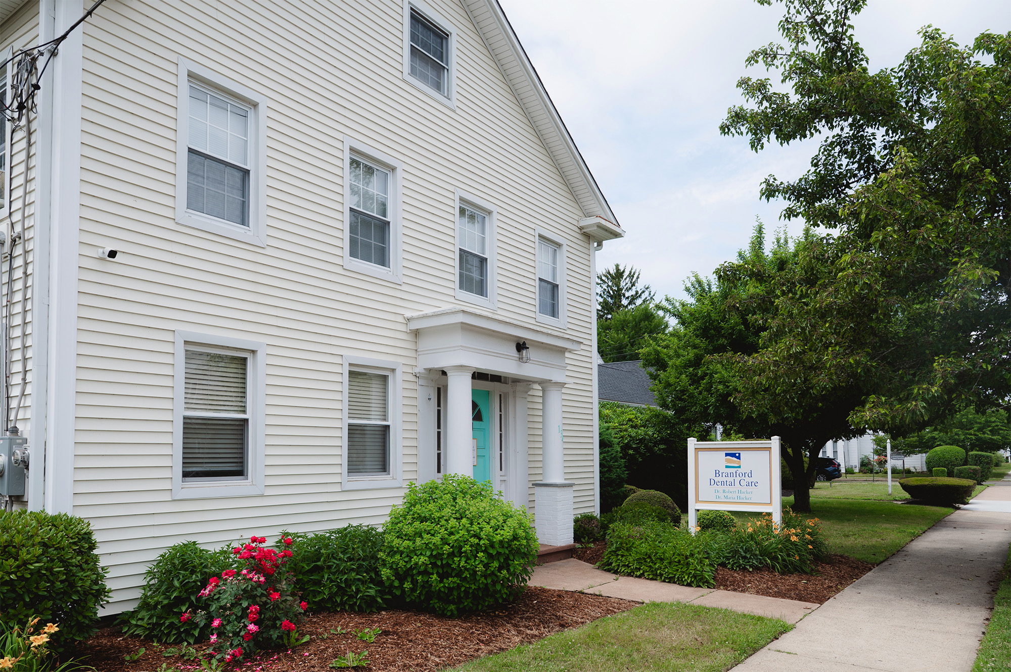 Exterior of Branford Dental Care building with white siding, teal door, and garden with flowers.