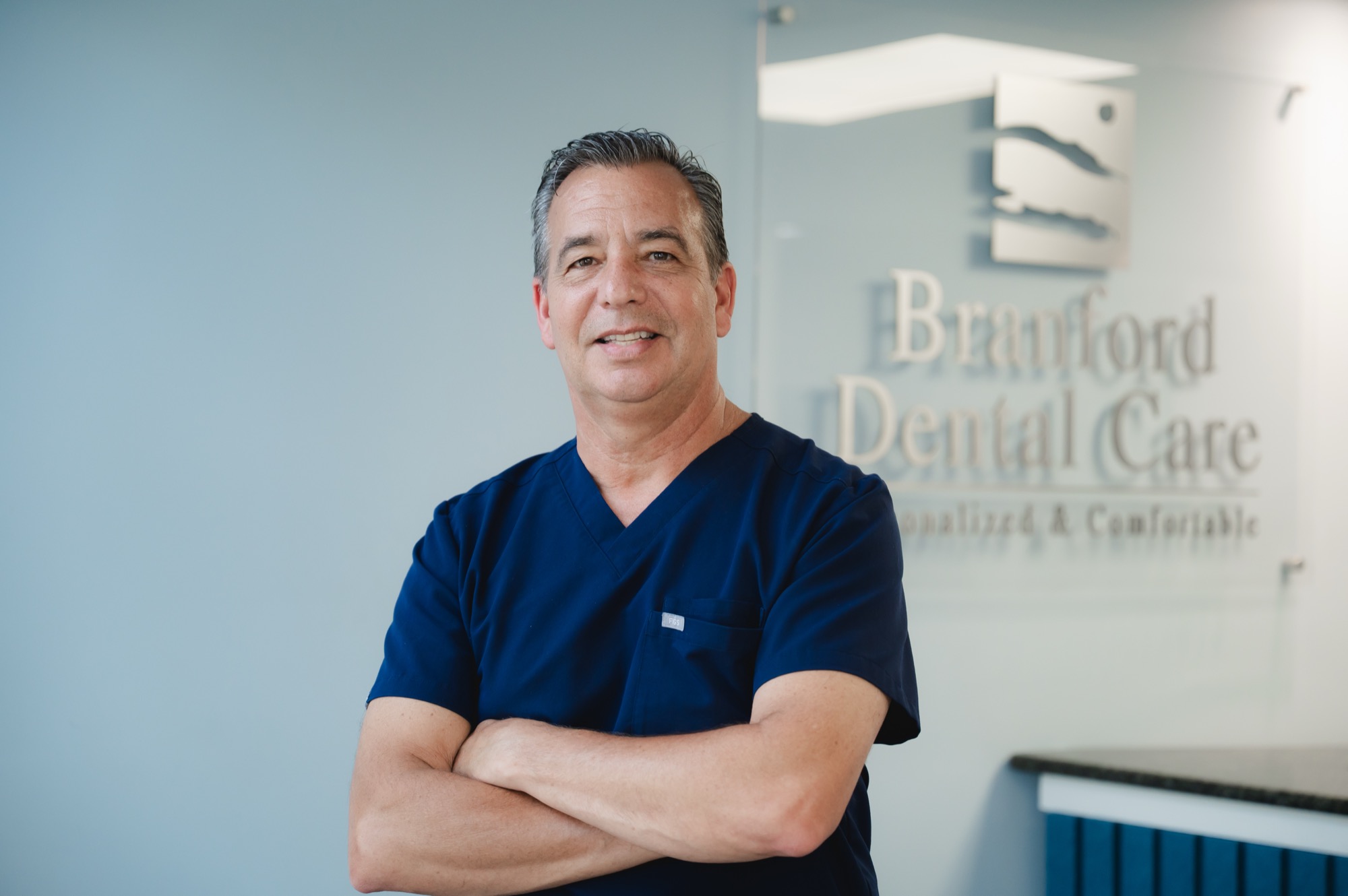 Smiling male dentist in navy scrubs standing with arms crossed in front of Branford Dental Care sign.
