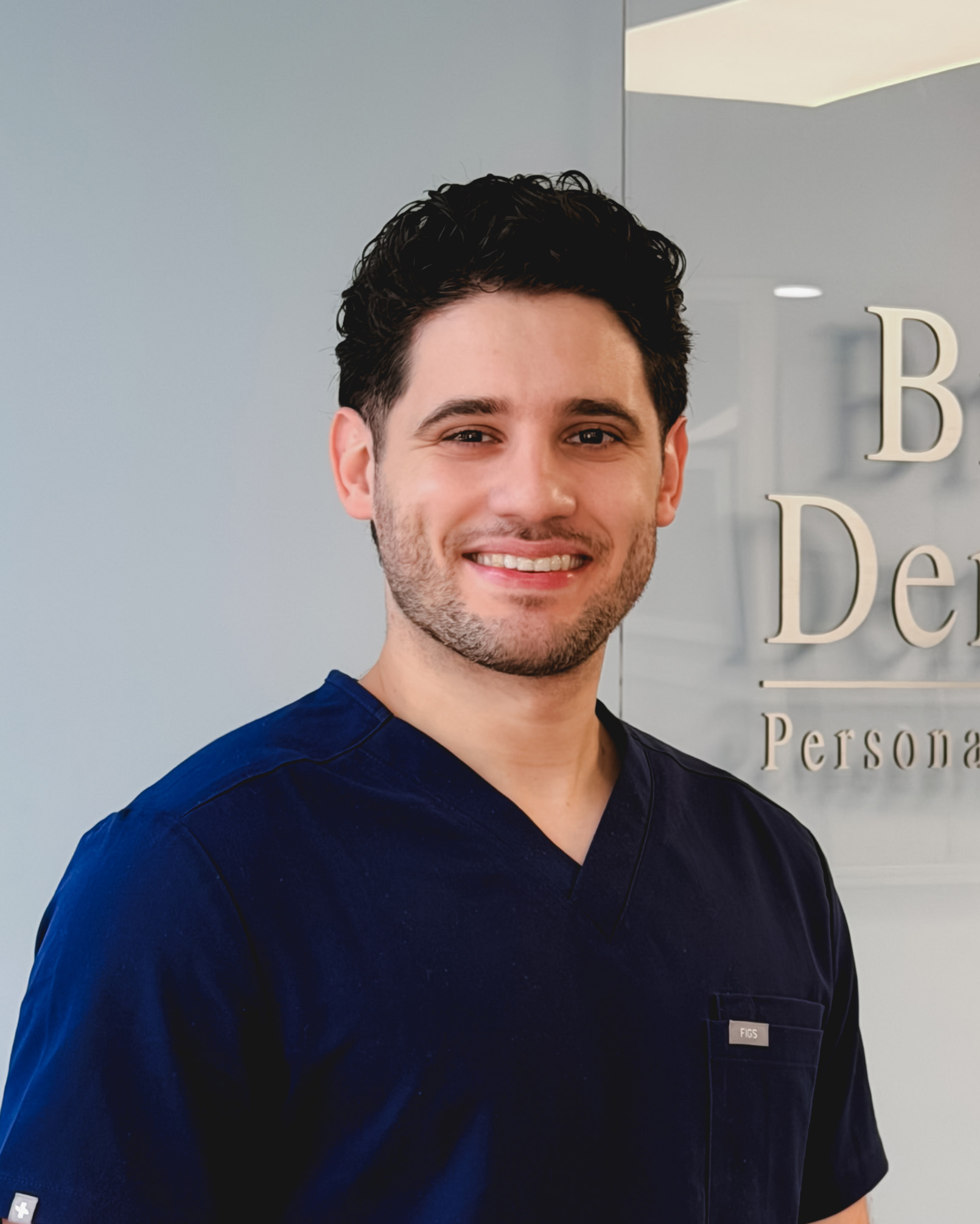 Smiling man with dark hair wearing navy blue scrubs in a dental office.