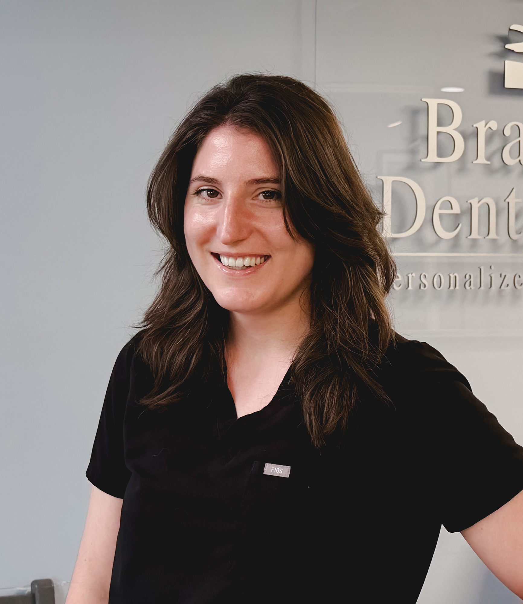 Smiling woman with dark hair wearing black scrubs stands in front of a dental office sign.