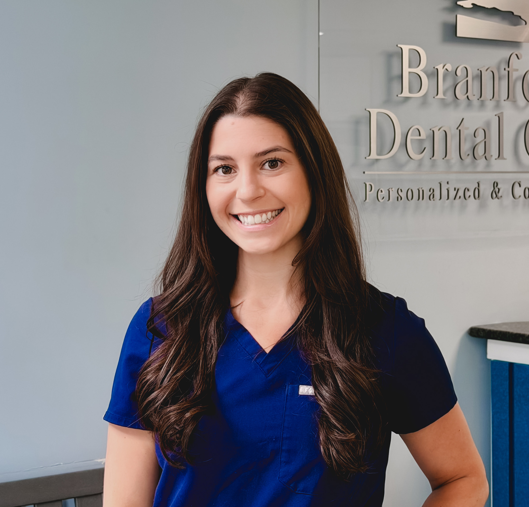 Smiling woman with long brown hair wearing blue dental scrubs standing in dental office.
