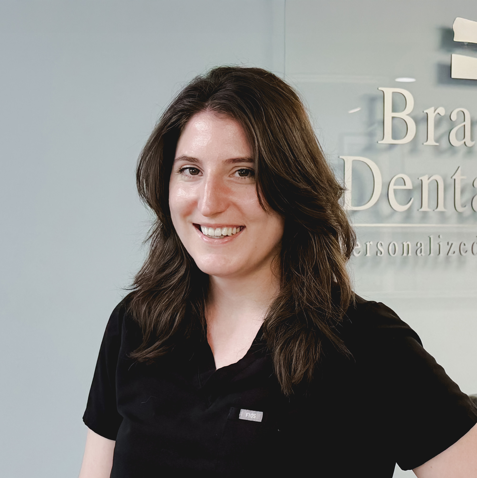 Smiling woman with shoulder-length brown hair wearing a black scrub top standing in front of a glass wall with partially visible text 'Branford Dental Care personalized'.