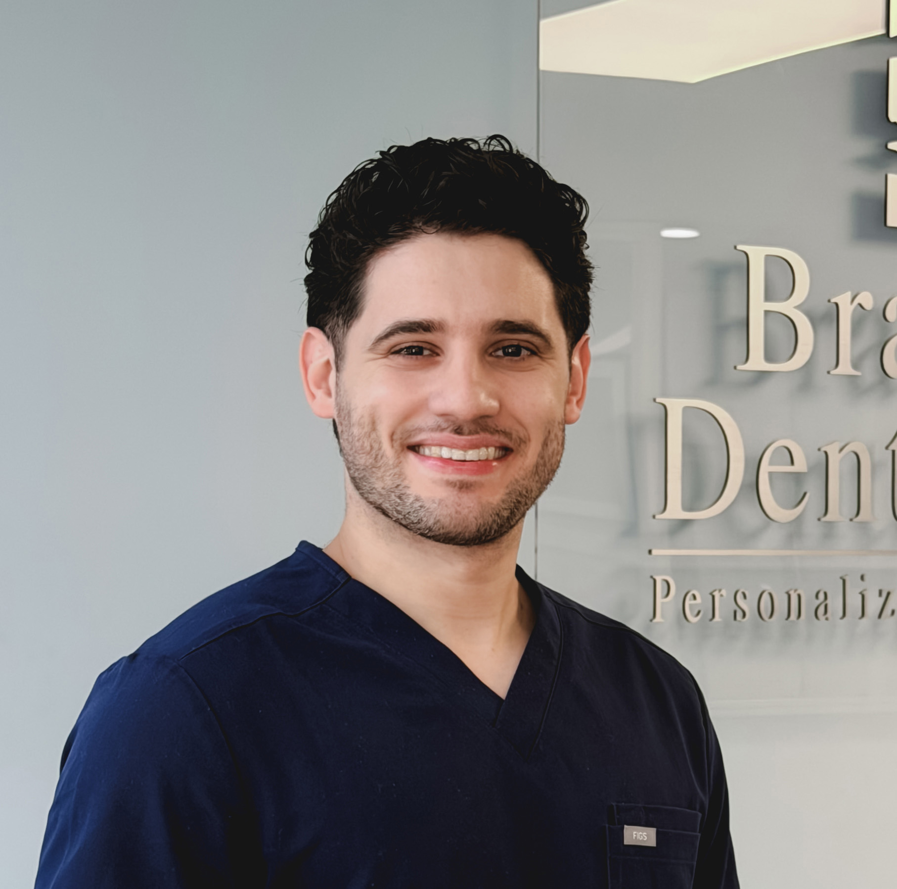 Smiling man with dark curly hair and beard wearing navy blue medical scrubs standing in front of a gray wall with partial dental care signage.