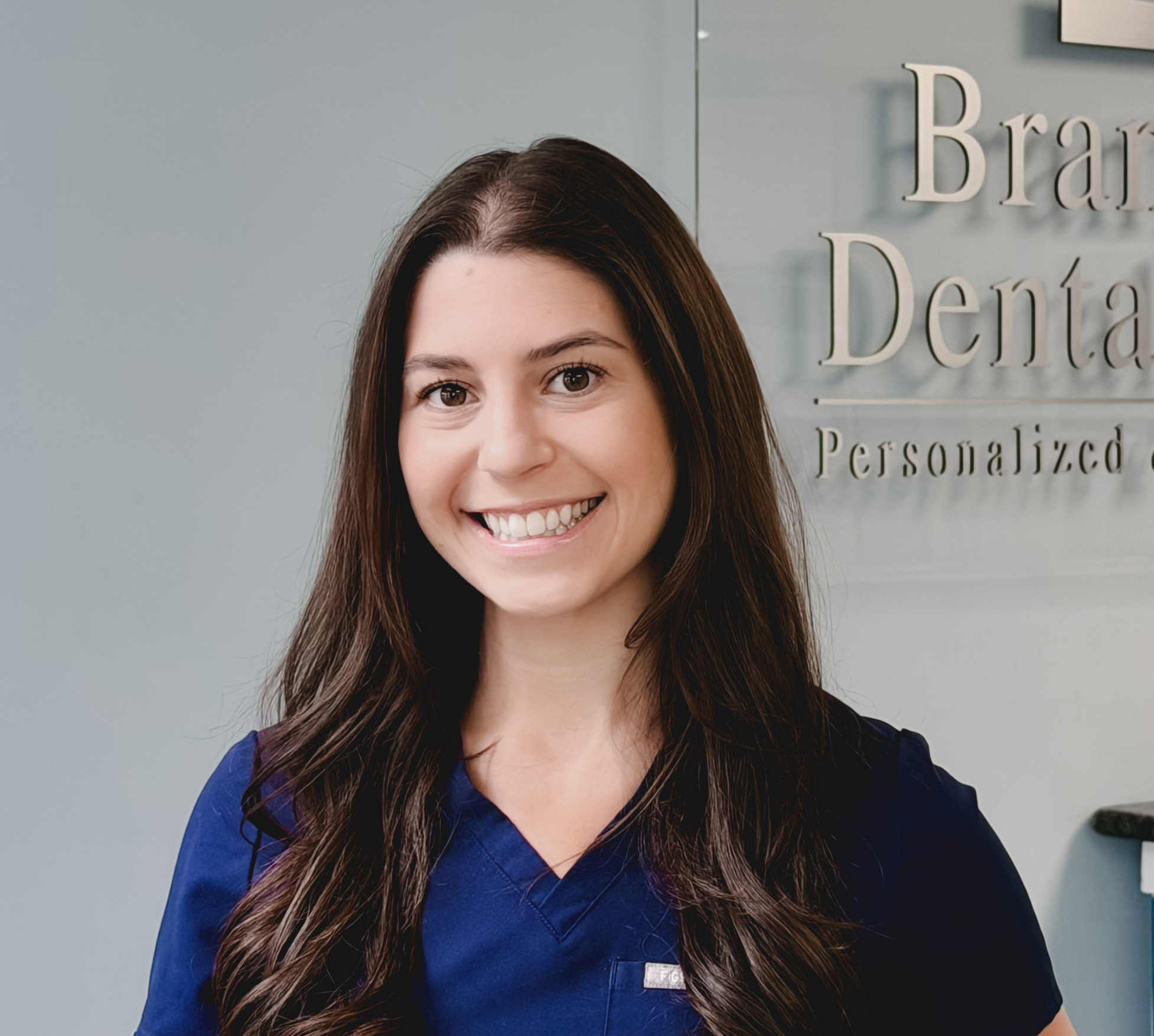 Smiling woman with long dark hair wearing navy blue scrubs standing indoors in front of a glass sign that partially reads 'Branford Dental Personalized'.