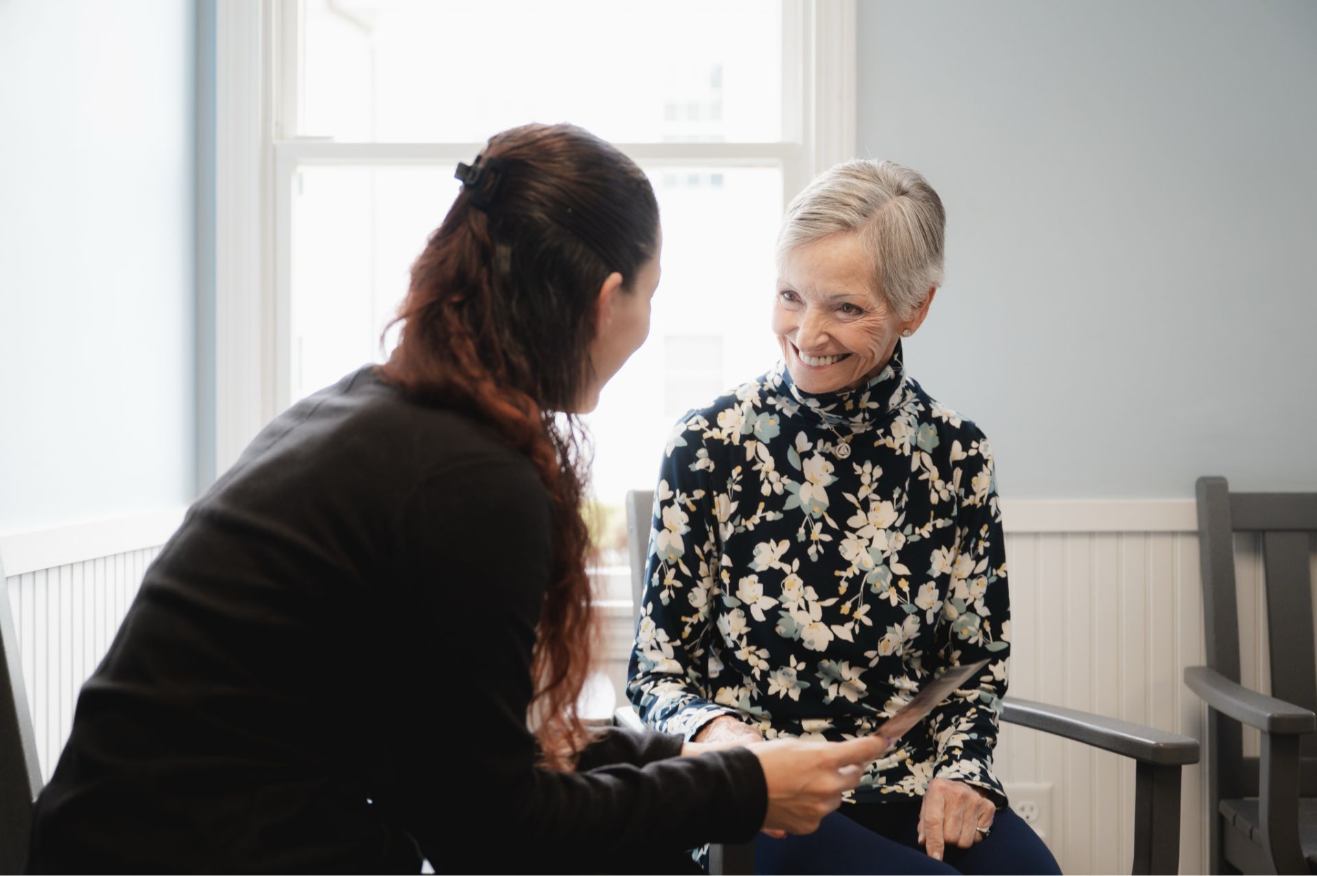 Smiling elderly woman in a floral turtleneck sitting and talking with a younger woman who holds a document in a bright room with light blue walls.