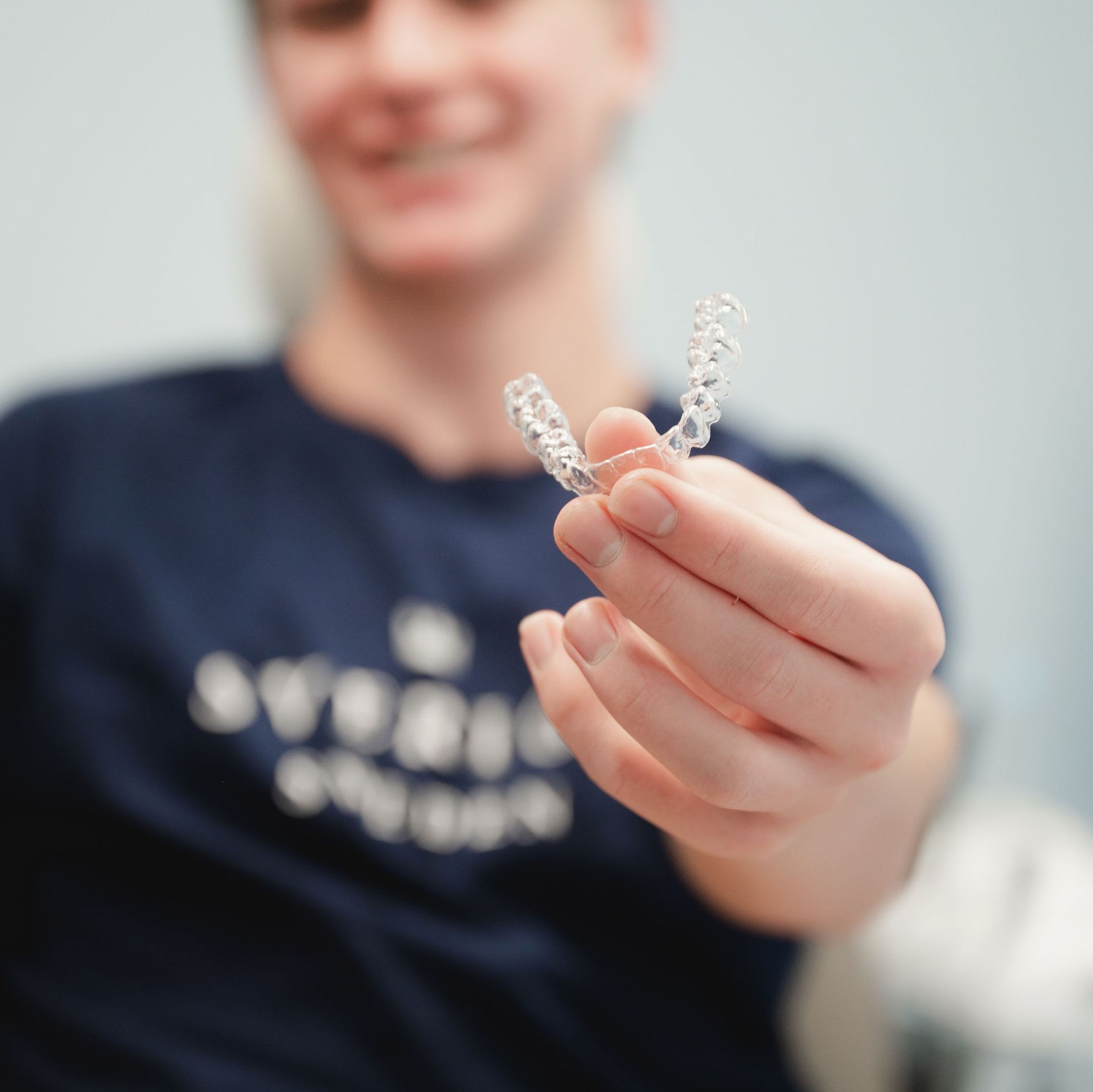 Person holding a clear dental aligner between their thumb and finger, smiling in the blurred background.