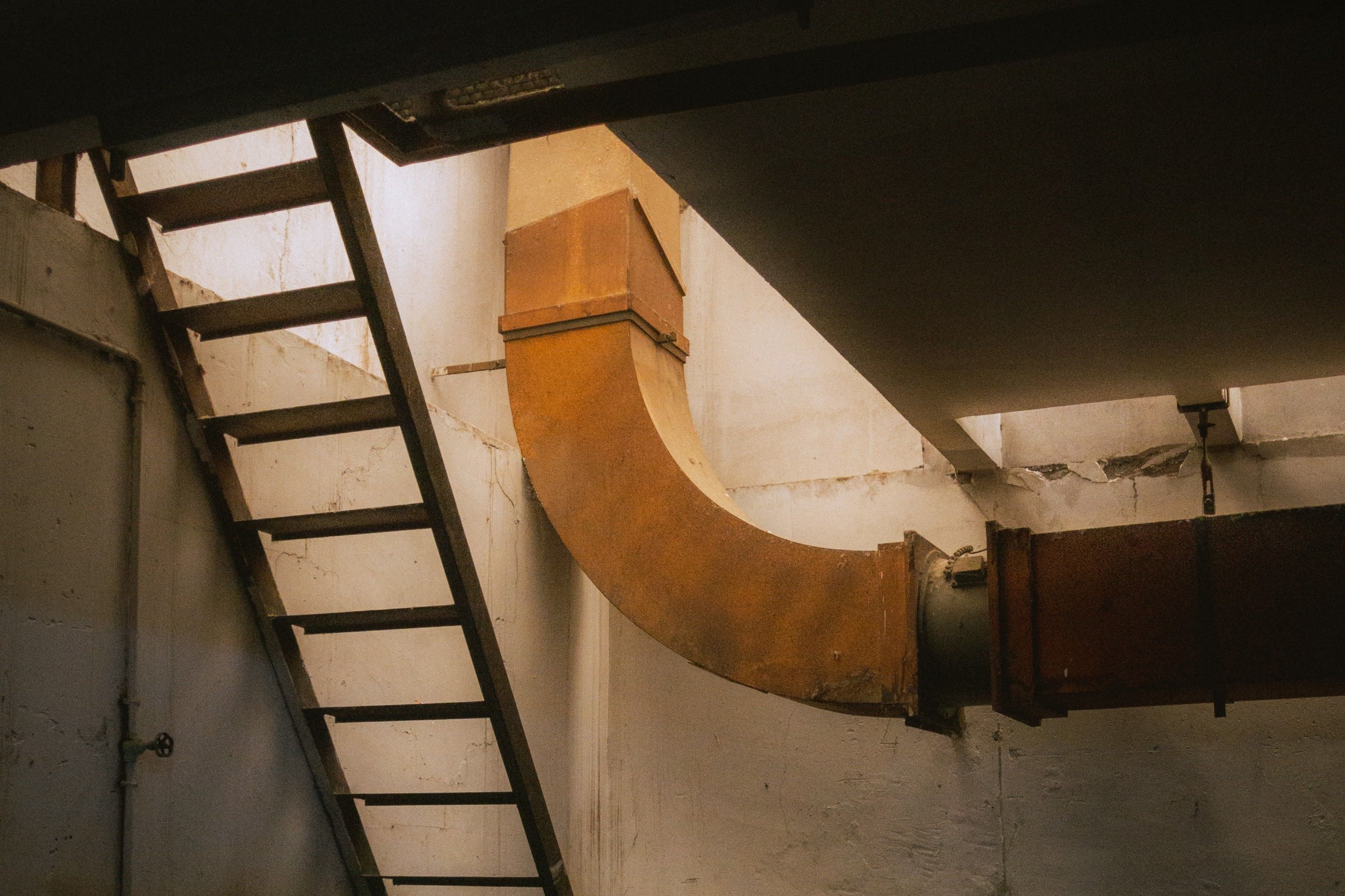 Rusted ductwork elbow turning across a basement plant room wall, with a steel access ladder in the foreground.