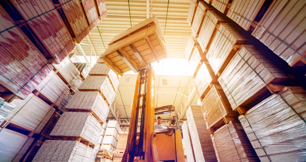 Worker operating forklift in warehouse stacking tall columns of large cardboard boxes.