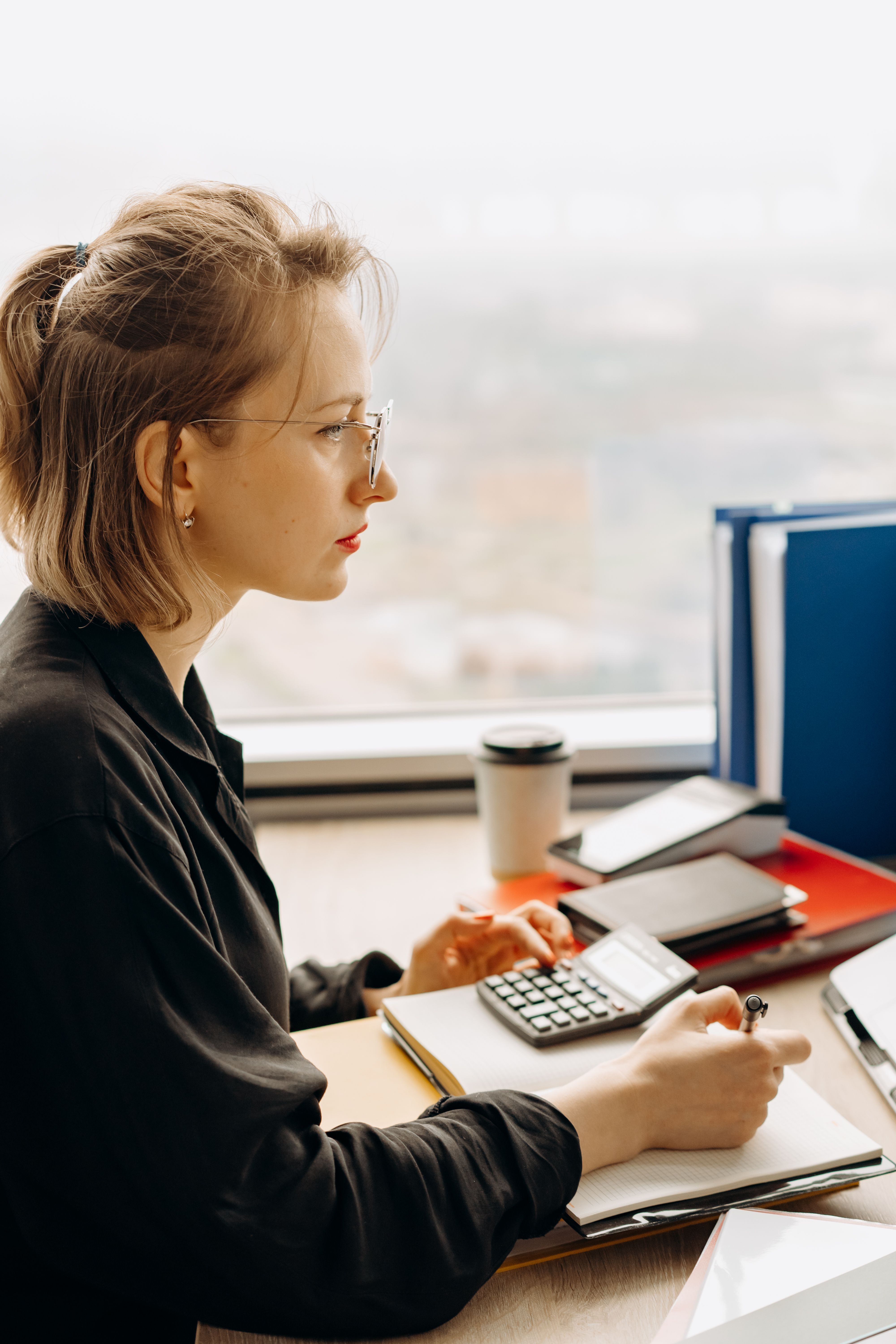 Young woman with glasses using calculator and writing in a notebook at a desk by a window.